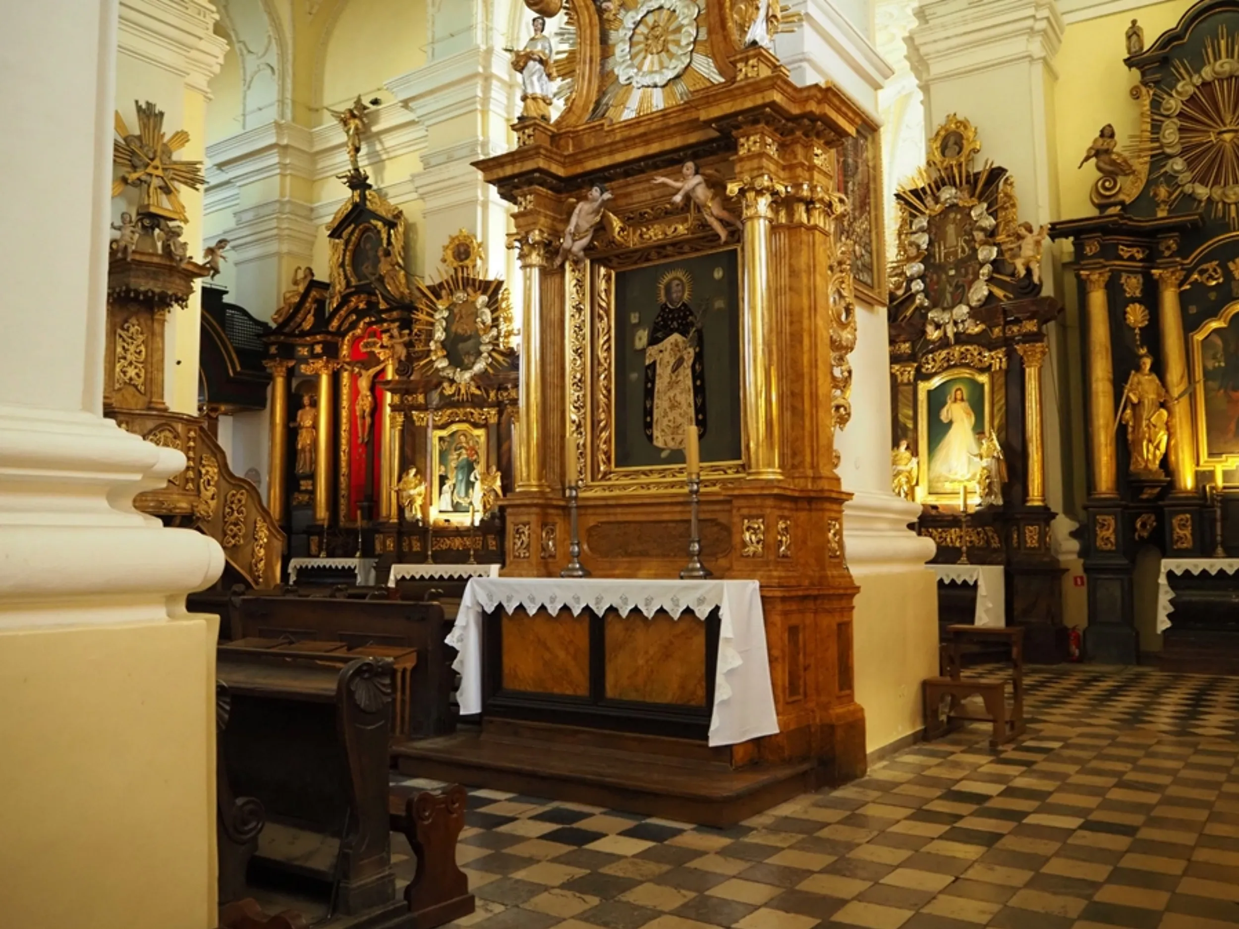 Wnętrze bazyliki z bocznym ołtarzem i bogato zdobionymi, barokowymi nastawami. Widoczne ławki, biało-czarna posadzka oraz obrazy religijne w złoconych ramach.