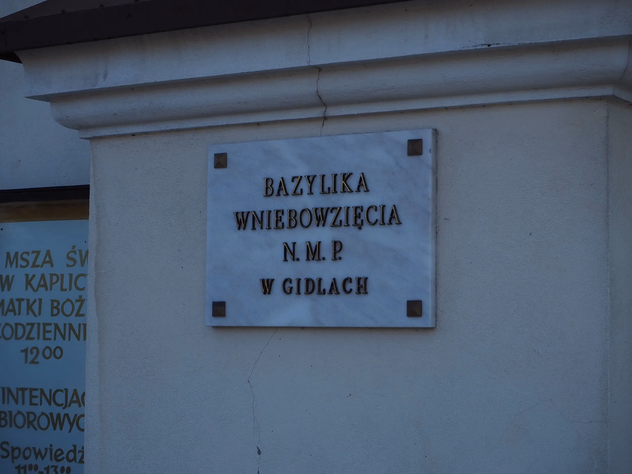 Tablica z napisem „Bazylika Wniebowzięcia N.M.P. w Gidlach” umieszczona na jasnej ścianie przy wejściu do świątyni.