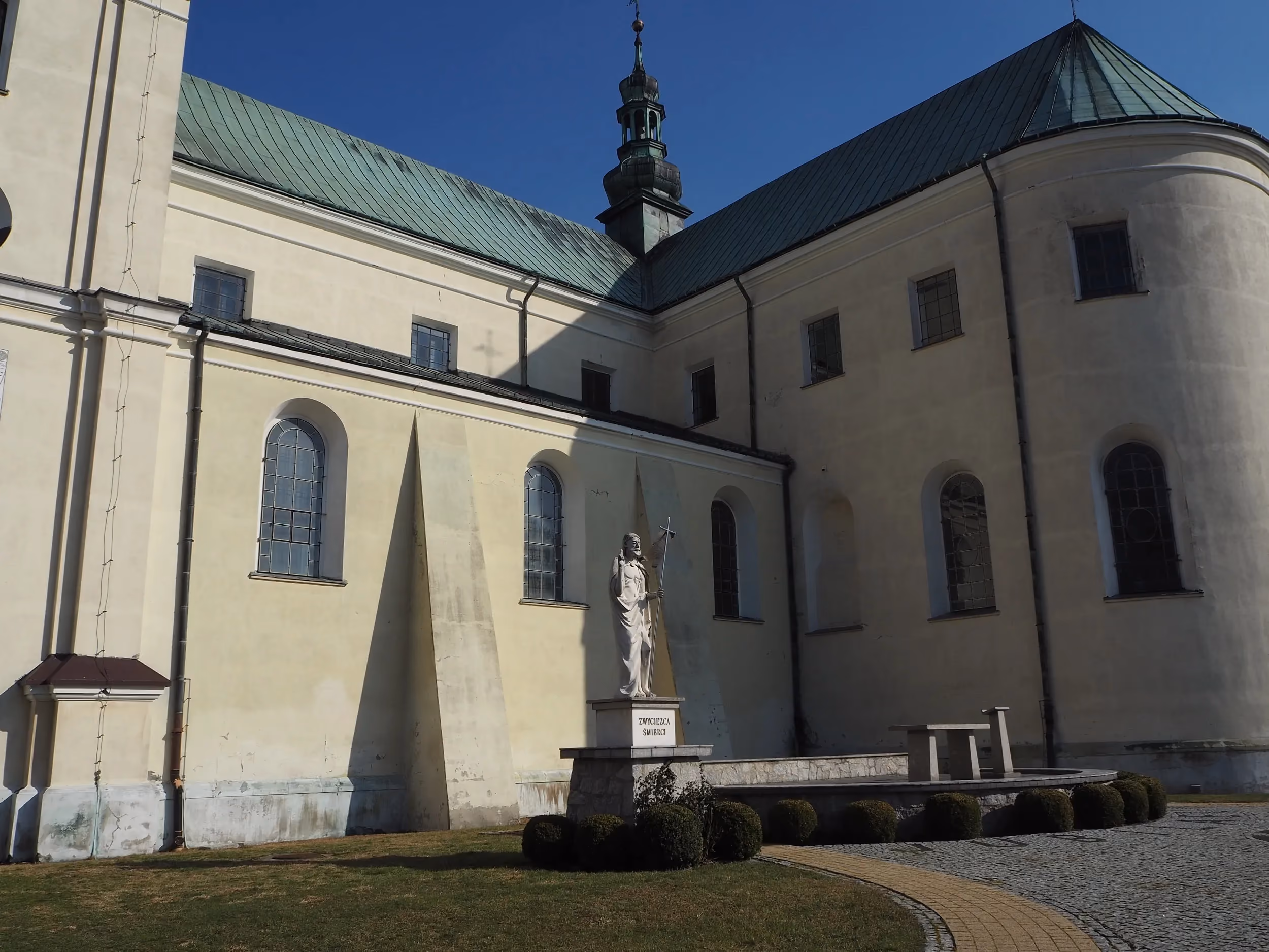 Boczna część Bazyliki Wniebowzięcia NMP w Gidlach. Na dziedzińcu widoczna figura Chrystusa Zmartwychwstałego oraz fragment zabudowań świątyni.