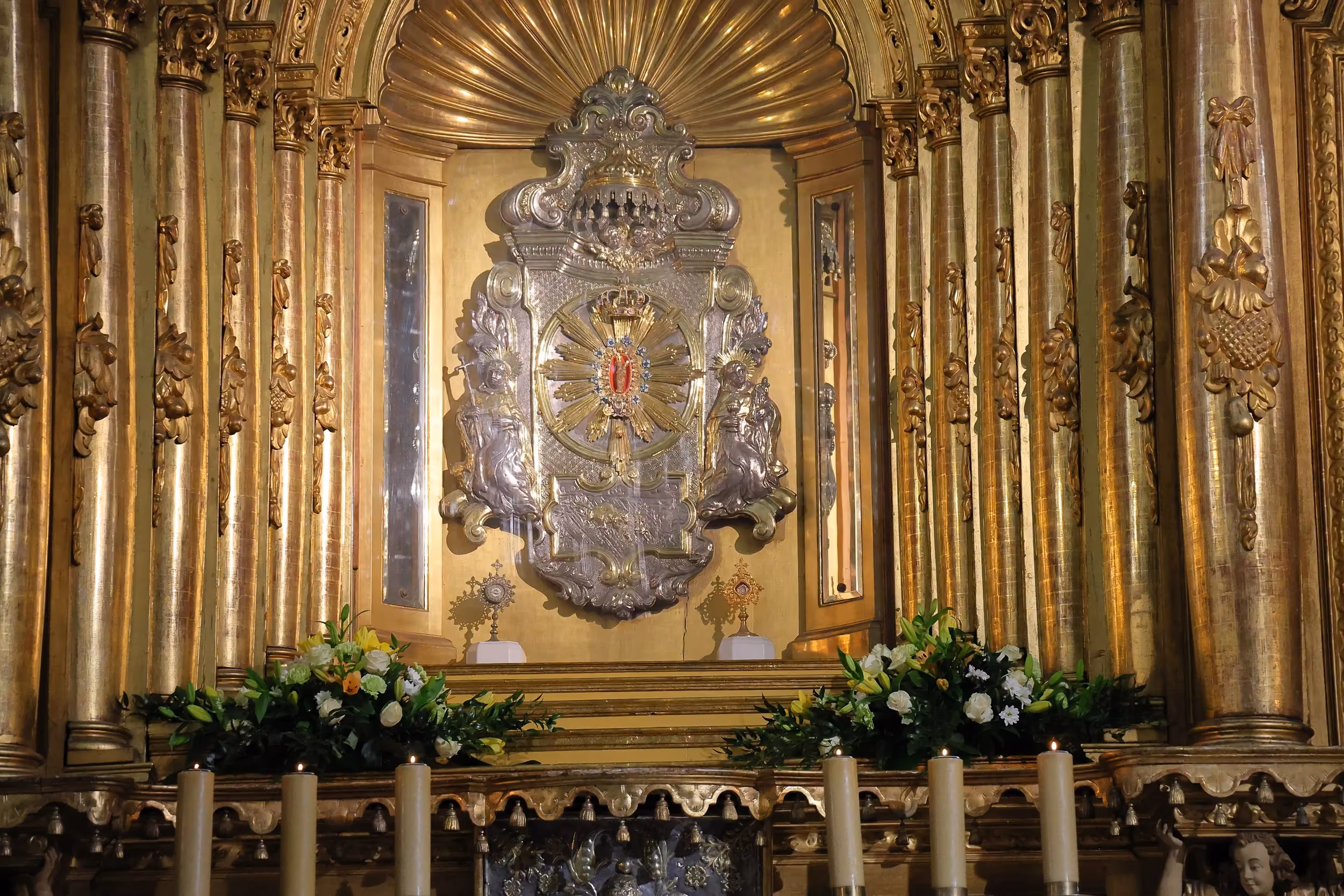 Fragment ołtarza głównego bazyliki z ozdobną nastawą, świecami i kompozycjami kwiatowymi ustawionymi przed centralnym miejscem kultu.