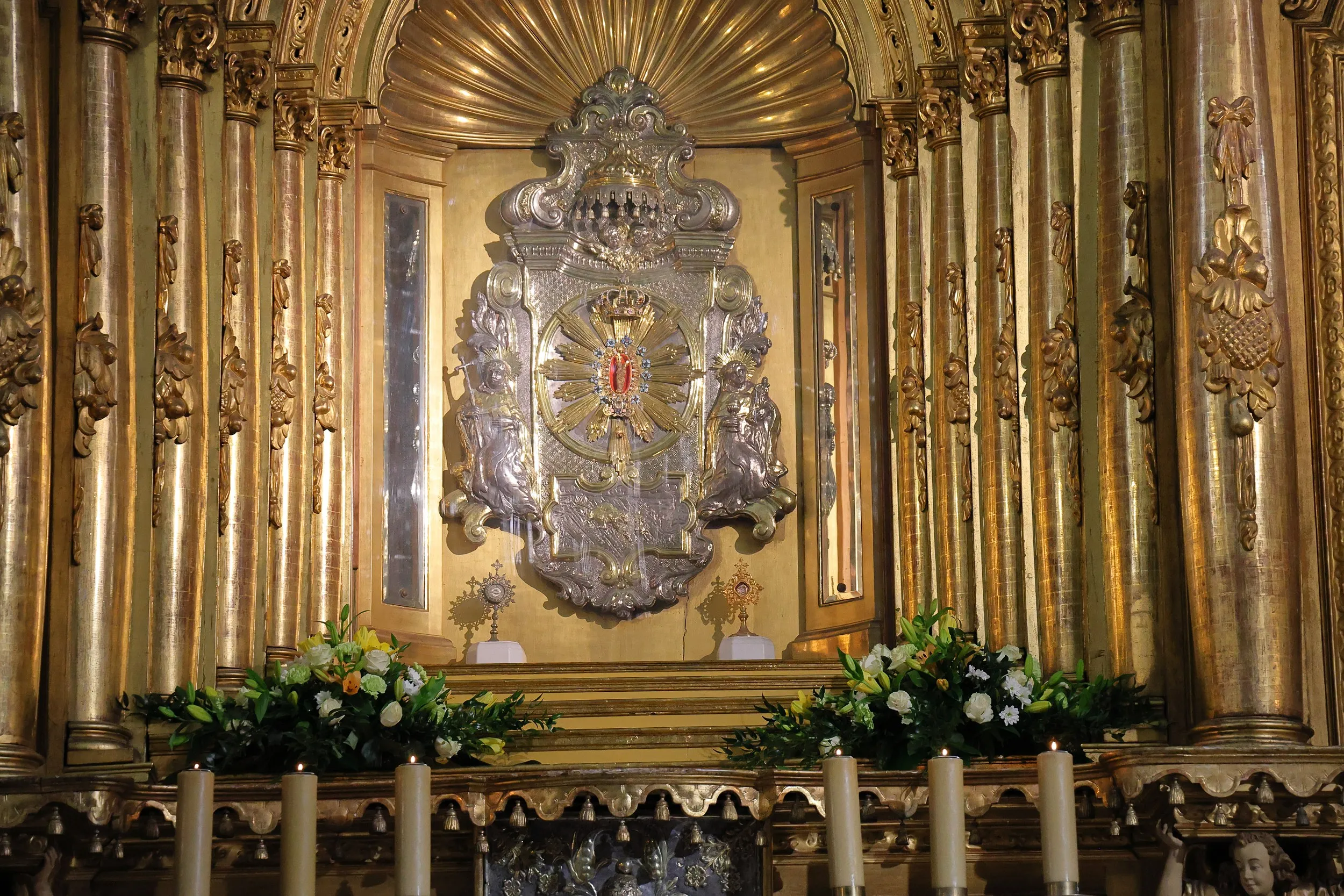 Fragment ołtarza głównego bazyliki z ozdobną nastawą, świecami i kompozycjami kwiatowymi ustawionymi przed centralnym miejscem kultu.