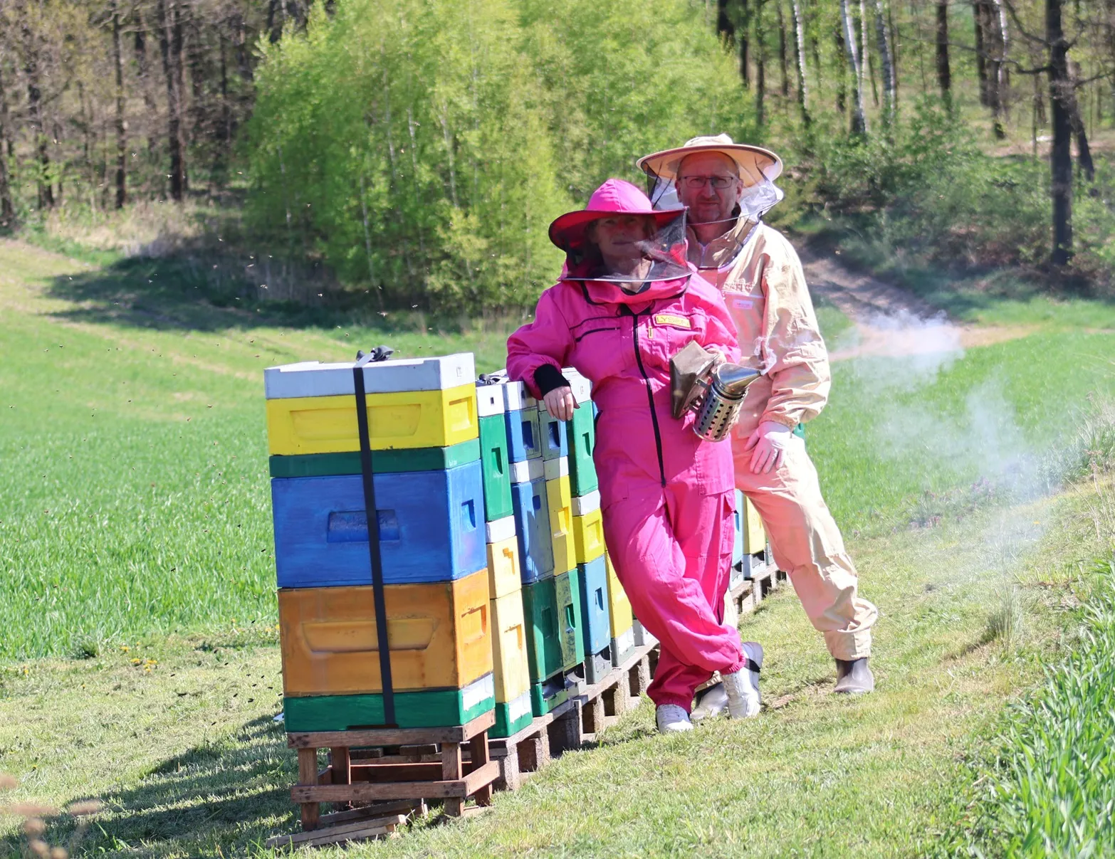 Dwoje pszczelarzy (kobieta w różowym kombinezonie, mężczyzna w beżowym) opiera się o kolorowe ule na łące, obok unosi się dym z podkurzacza.
