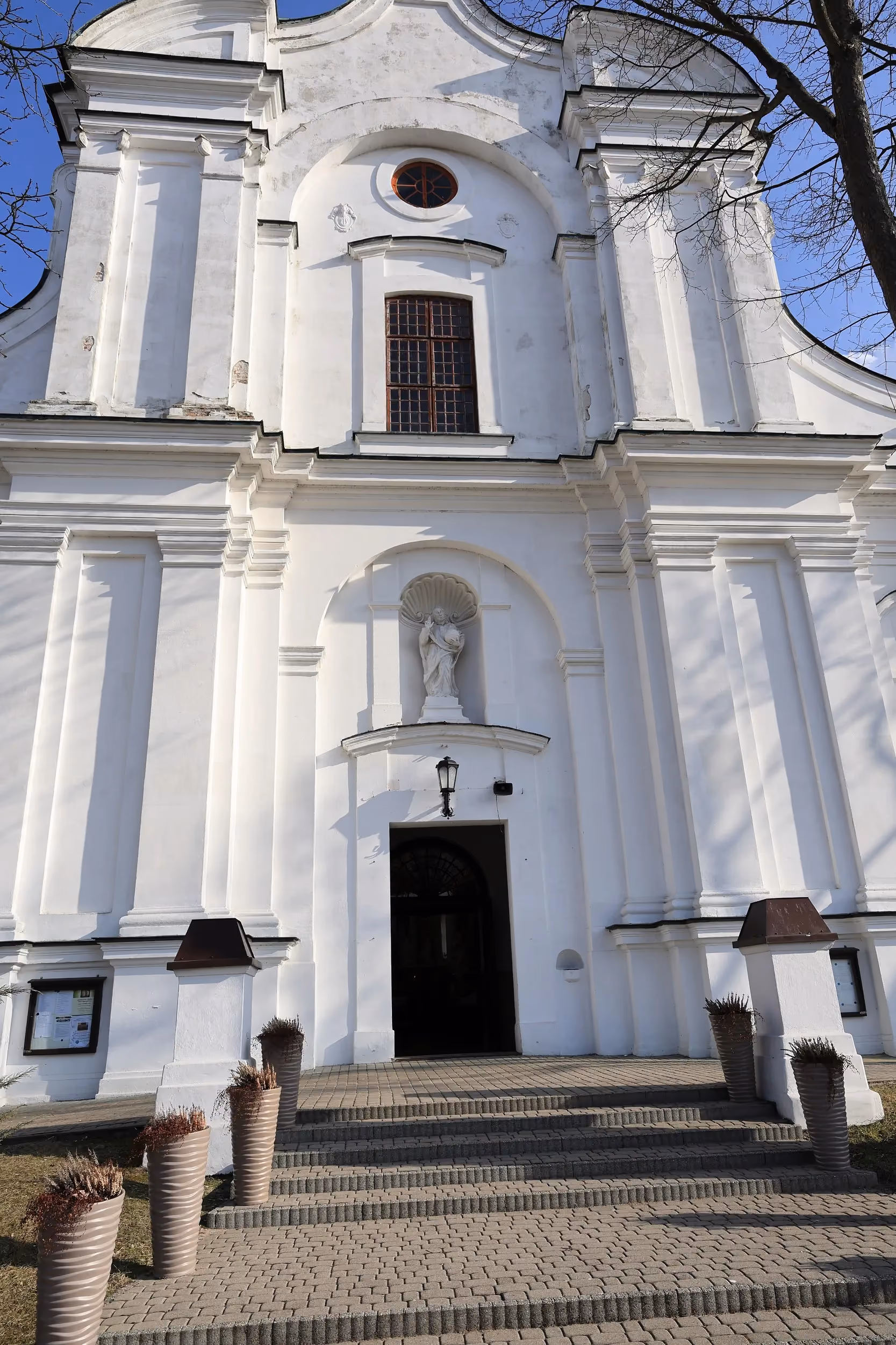 Frontowa fasada kościoła z głównym wejściem, schodami i figurą umieszczoną nad portalem. Budynek utrzymany jest w jasnej kolorystyce.