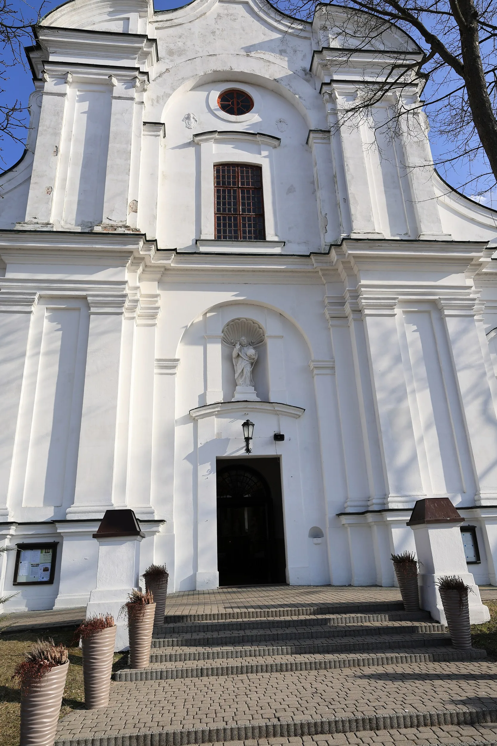 Frontowa fasada kościoła z głównym wejściem, schodami i figurą umieszczoną nad portalem. Budynek utrzymany jest w jasnej kolorystyce.