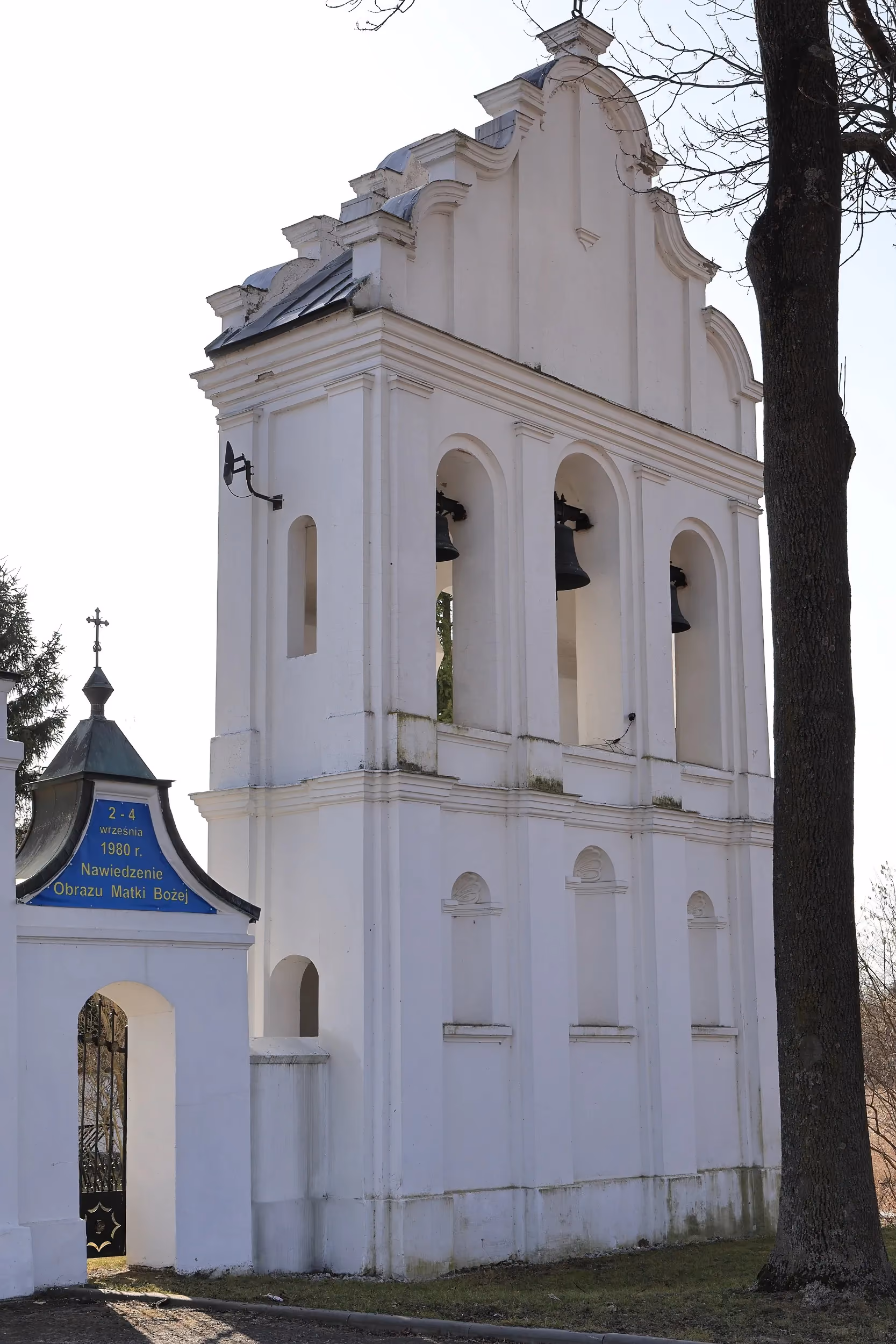 Biała dzwonnica z trzema dzwonami stojąca obok kościoła. Na pierwszym planie widoczne są drzewa i fragment ogrodzenia.