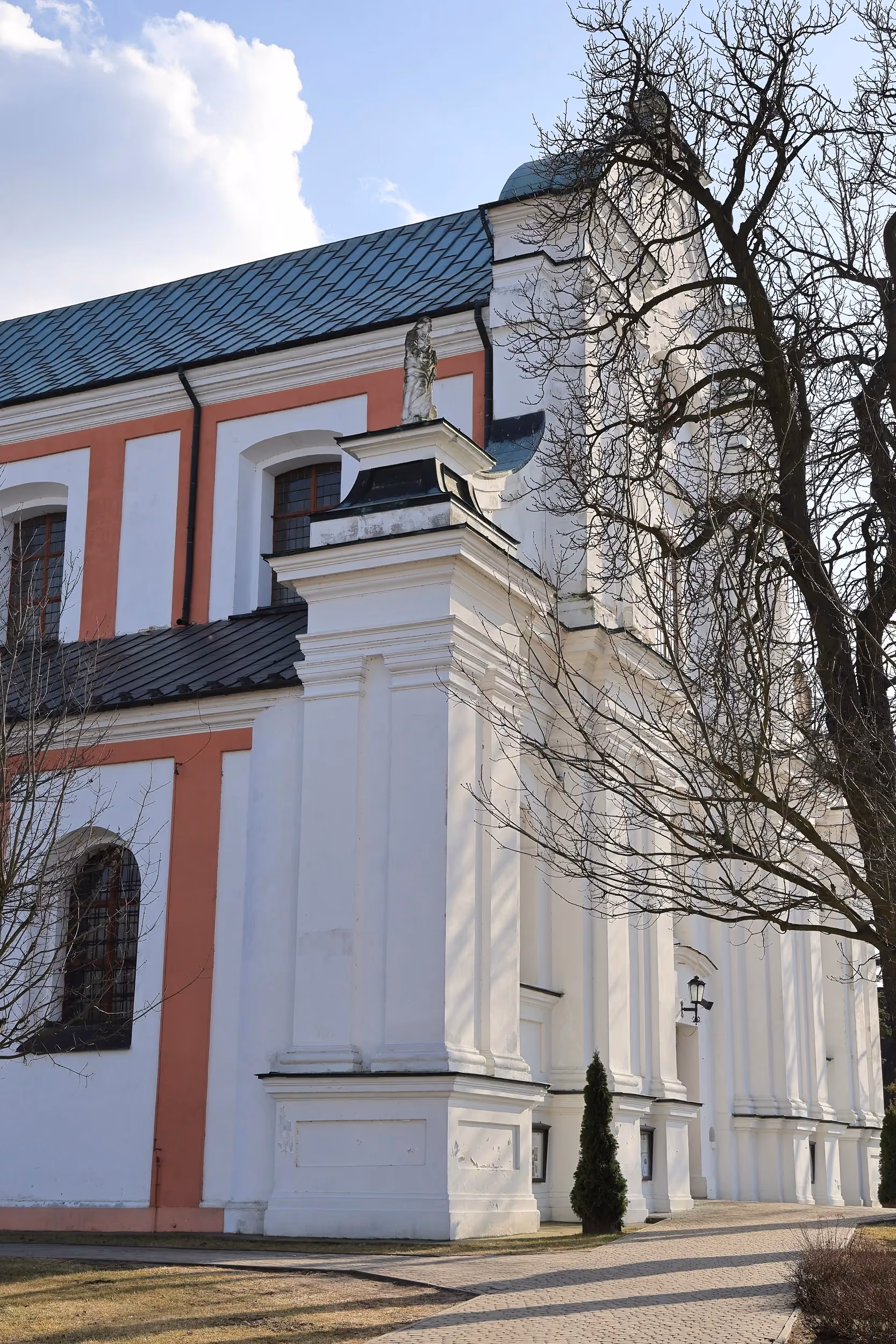 Boczna elewacja kościoła z białymi i różowymi elementami architektonicznymi. Do wejścia prowadzi brukowana alejka otoczona trawnikiem.