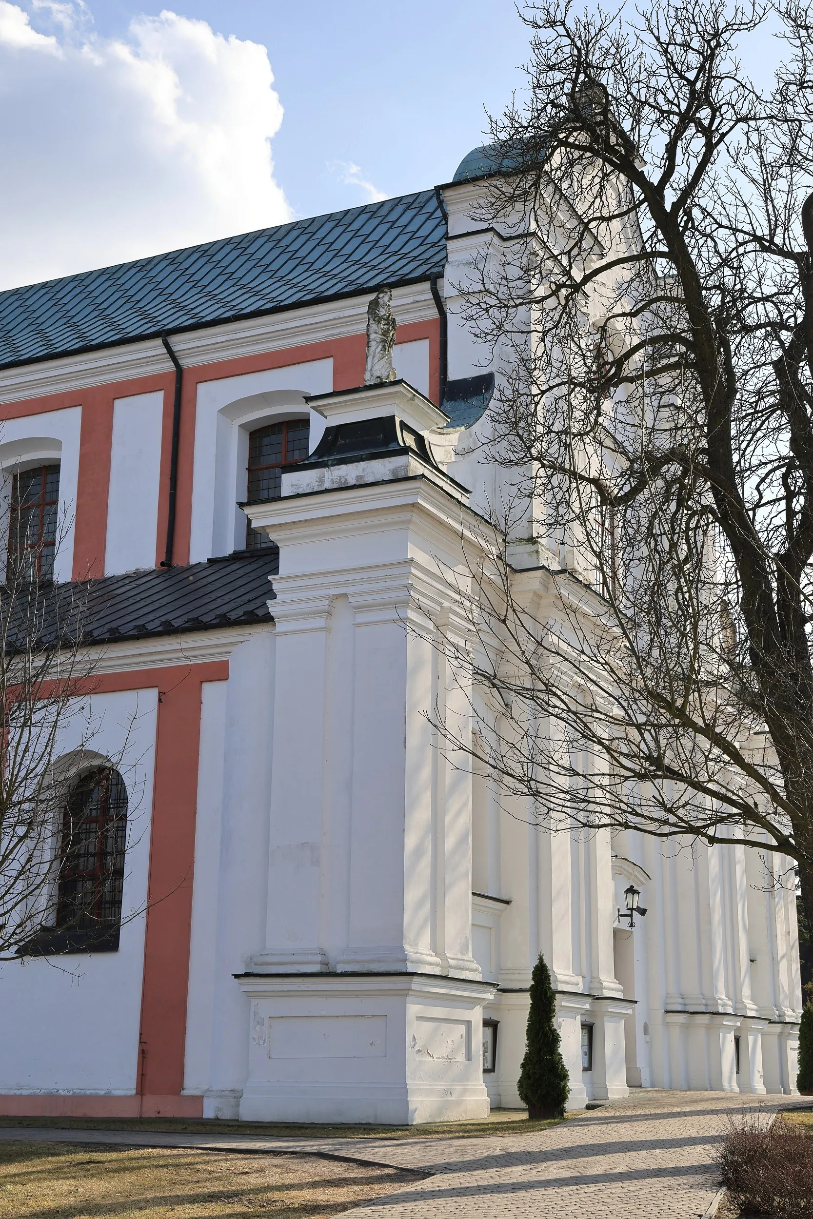 Boczna elewacja kościoła z białymi i różowymi elementami architektonicznymi. Do wejścia prowadzi brukowana alejka otoczona trawnikiem.