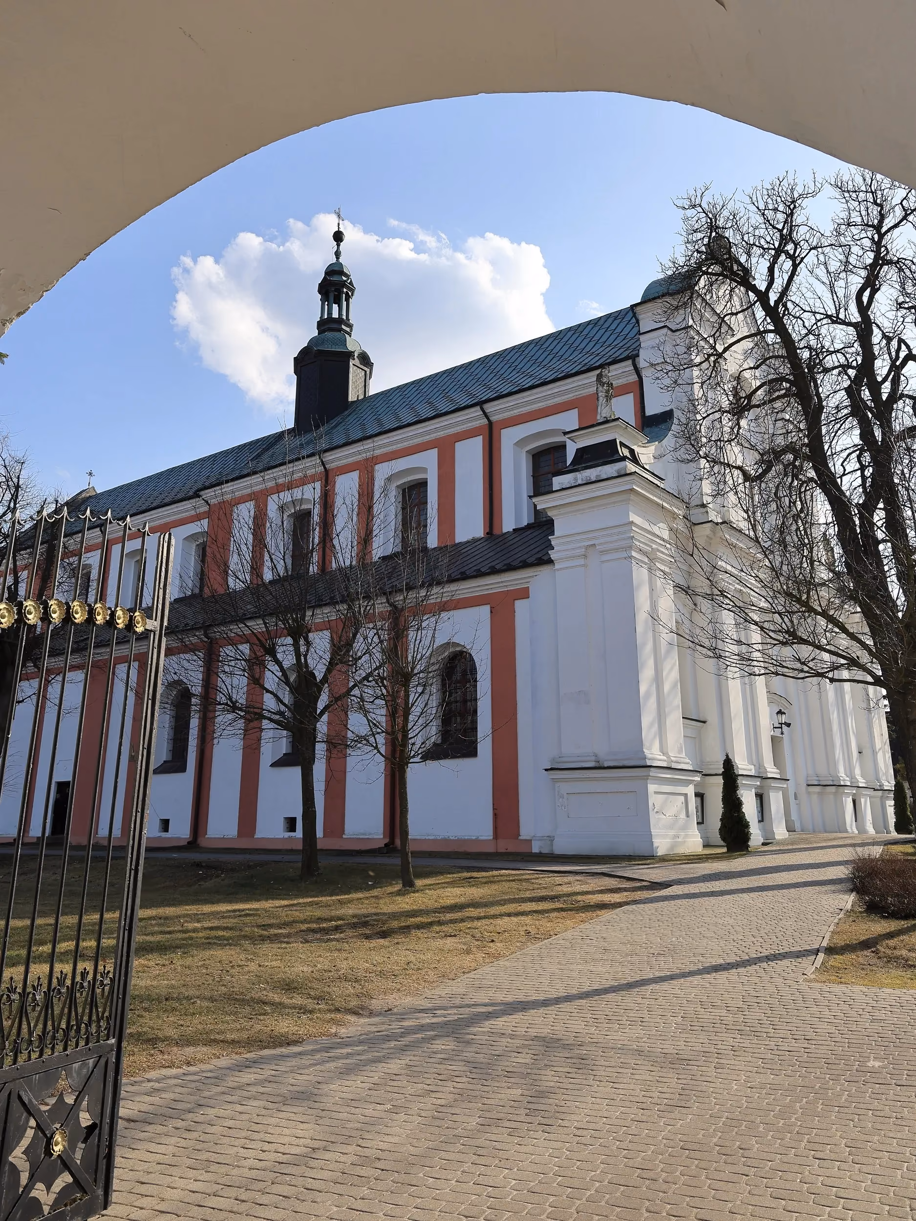 Boczna elewacja kościoła z białymi i różowymi elementami architektonicznymi. Do wejścia prowadzi brukowana alejka otoczona trawnikiem.