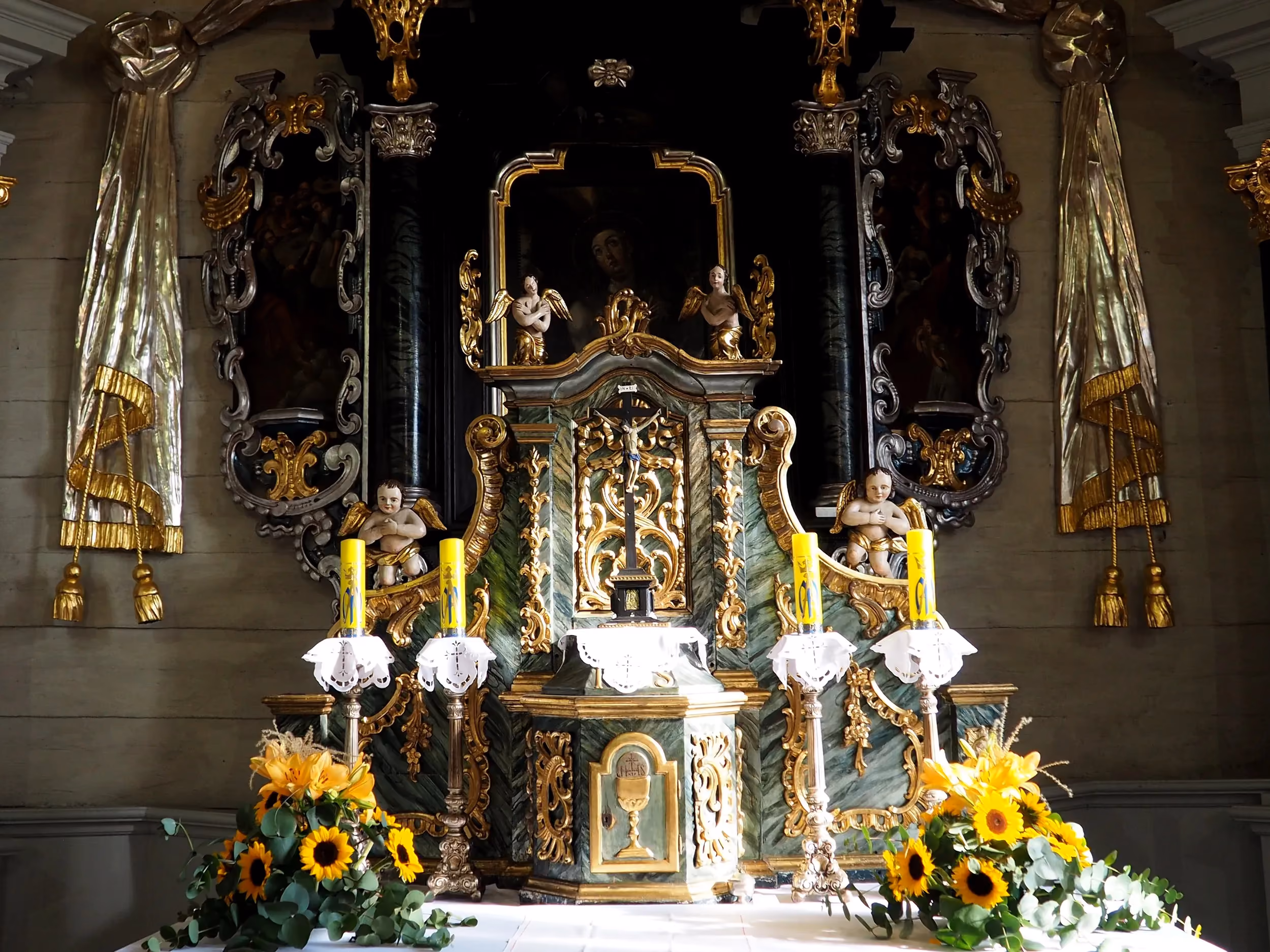 Zbliżenie na bogato zdobiony ołtarz główny z figurami aniołów, świecami i kompozycjami kwiatowymi. Tło stanowią ciemne elementy dekoracyjne.