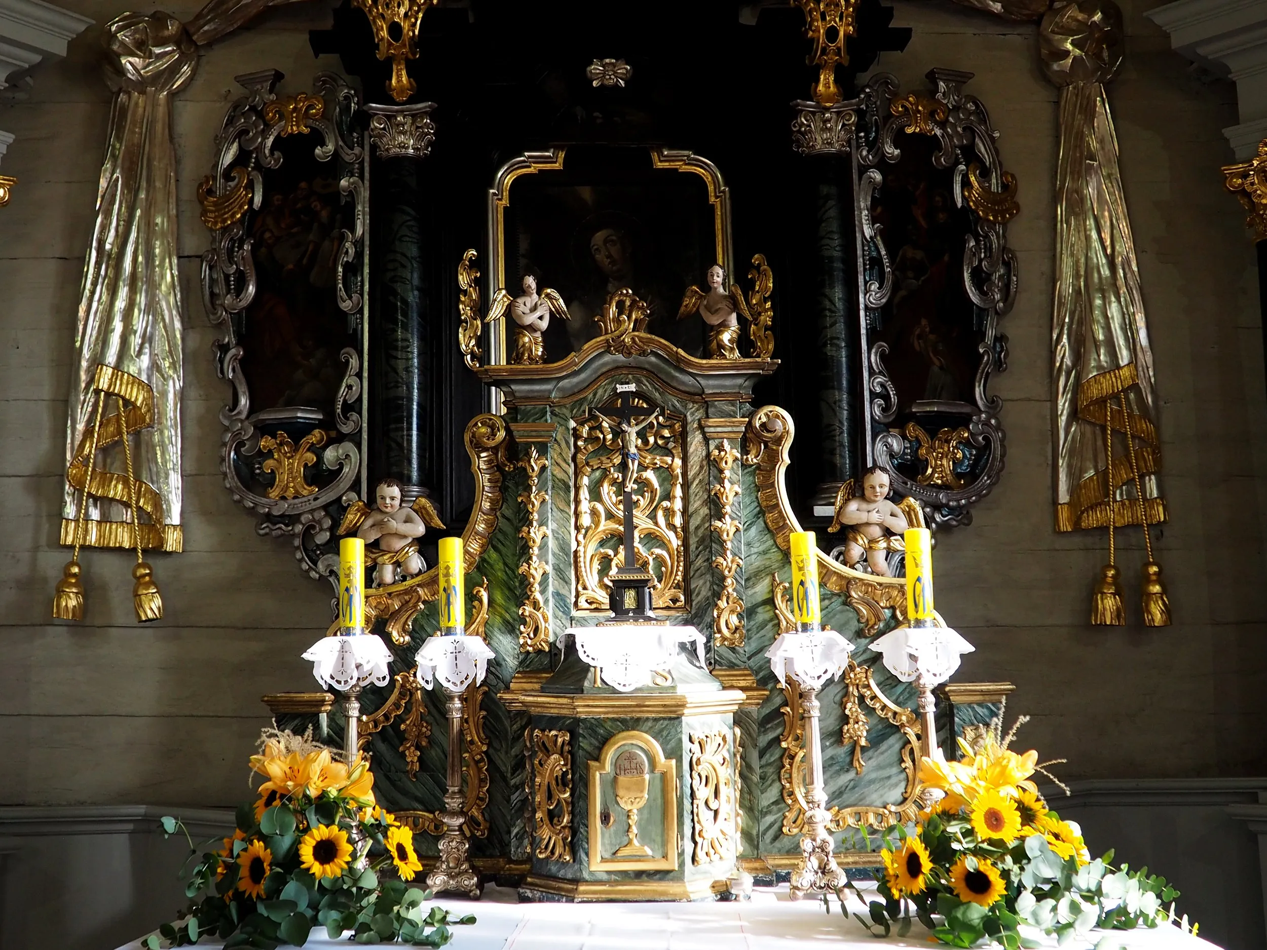 Zbliżenie na bogato zdobiony ołtarz główny z figurami aniołów, świecami i kompozycjami kwiatowymi. Tło stanowią ciemne elementy dekoracyjne.