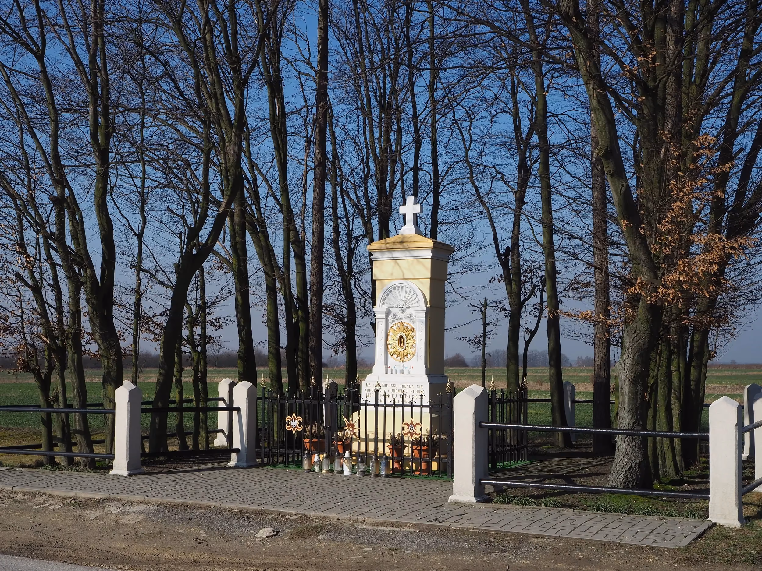 Kapliczka Matki Bożej Gidelskiej otoczona ogrodzeniem i drzewami. W tle rozciągają się pola, a przed kapliczką ustawiono znicze i kwiaty.