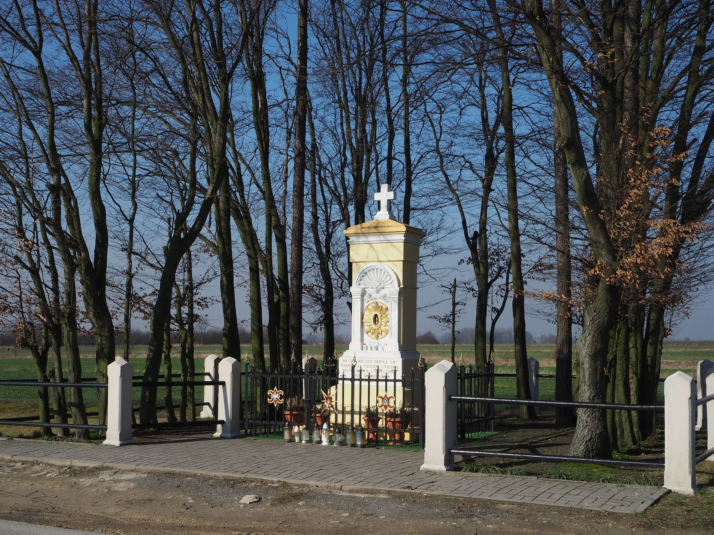 Kapliczka Matki Bożej Gidelskiej otoczona ogrodzeniem i drzewami. W tle rozciągają się pola, a przed kapliczką ustawiono znicze i kwiaty.