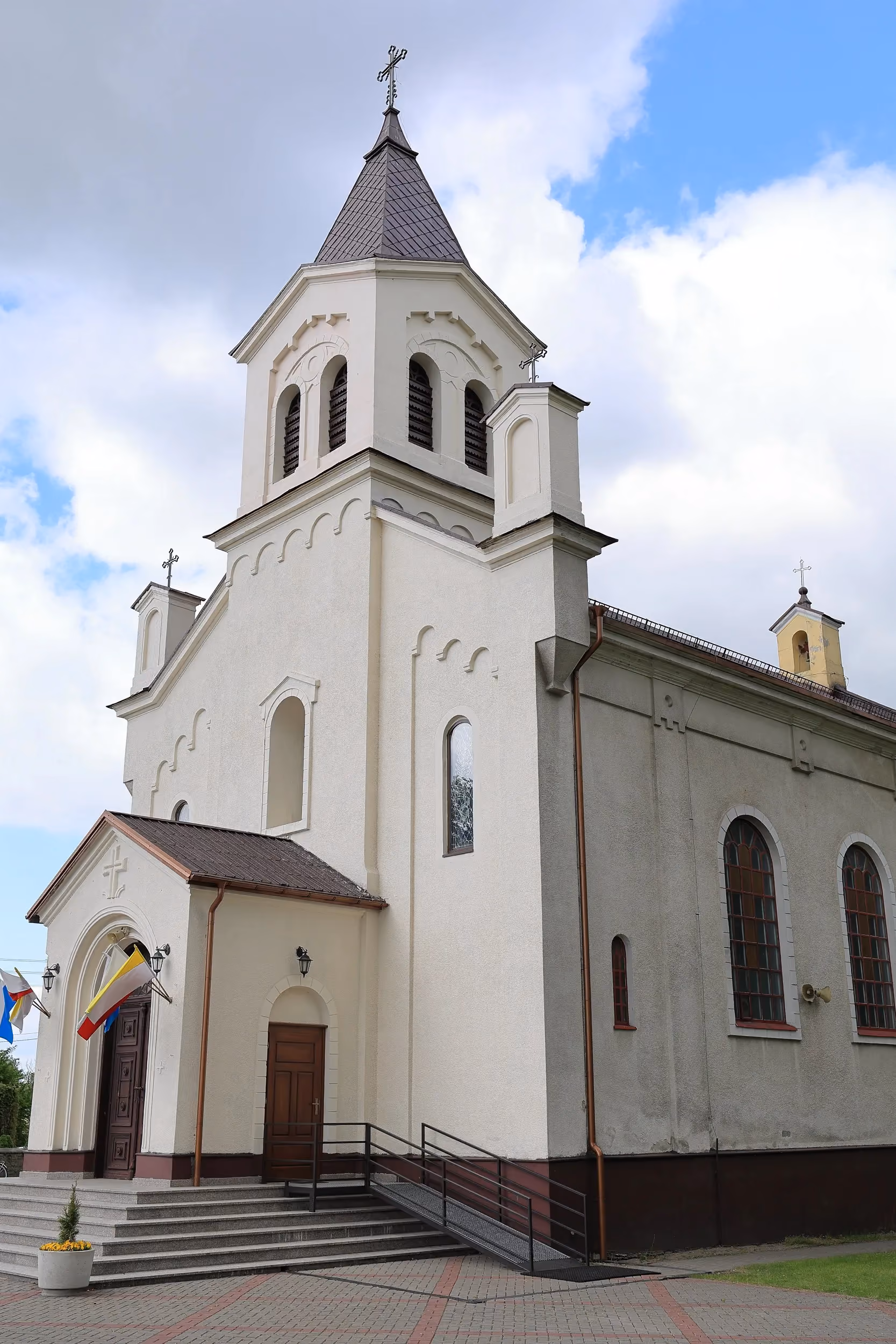 Widok zewnętrzny kościoła w Pławnie z jasną elewacją i wieżą zwieńczoną krzyżem. Do wejścia prowadzą schody oraz pochylnia dla osób z niepełnosprawnościami.