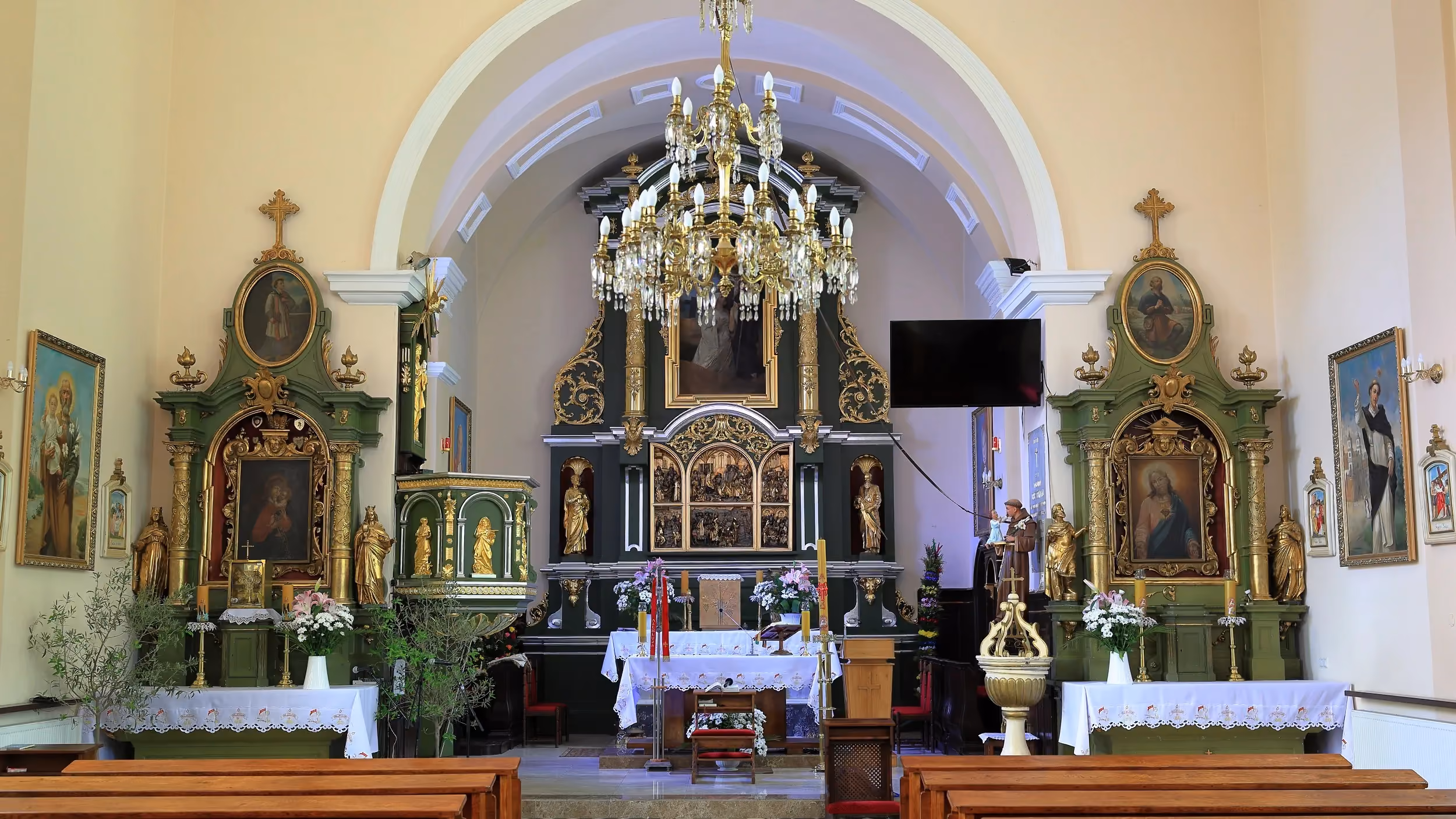 Wnętrze kościoła z głównym ołtarzem, bocznymi ołtarzami i żyrandolem. Przed ołtarzem ustawiono ławki, kwiaty i elementy liturgiczne.