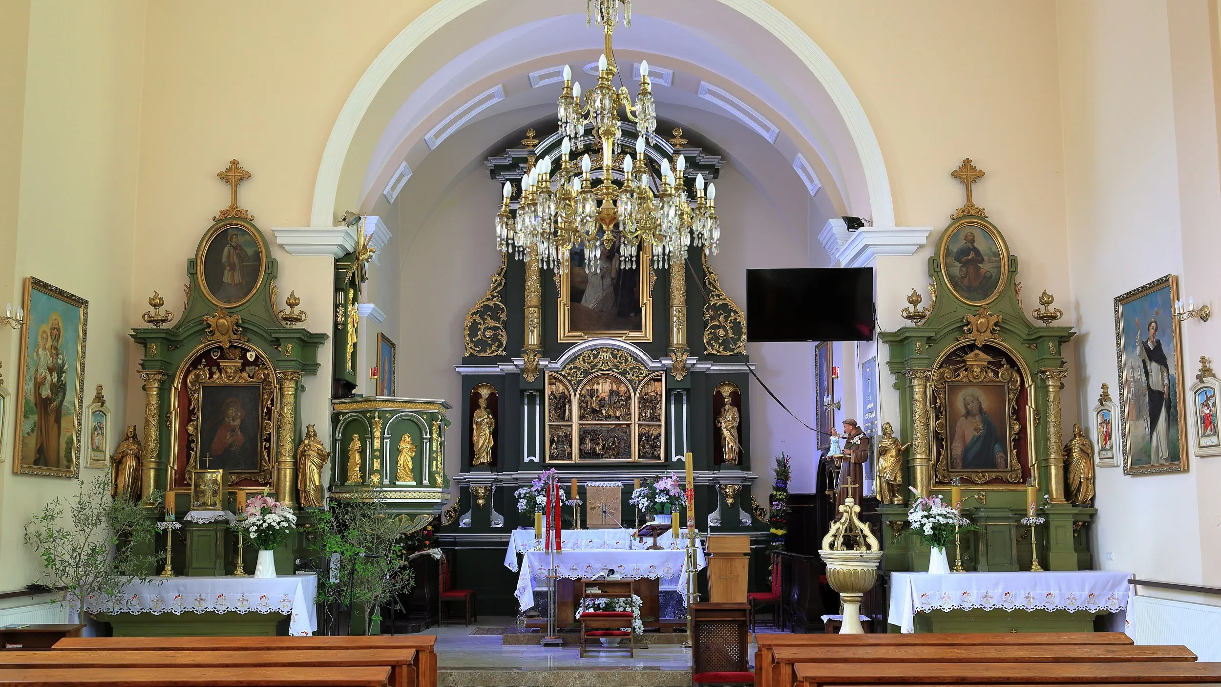 Wnętrze kościoła z głównym ołtarzem, bocznymi ołtarzami i żyrandolem. Przed ołtarzem ustawiono ławki, kwiaty i elementy liturgiczne.