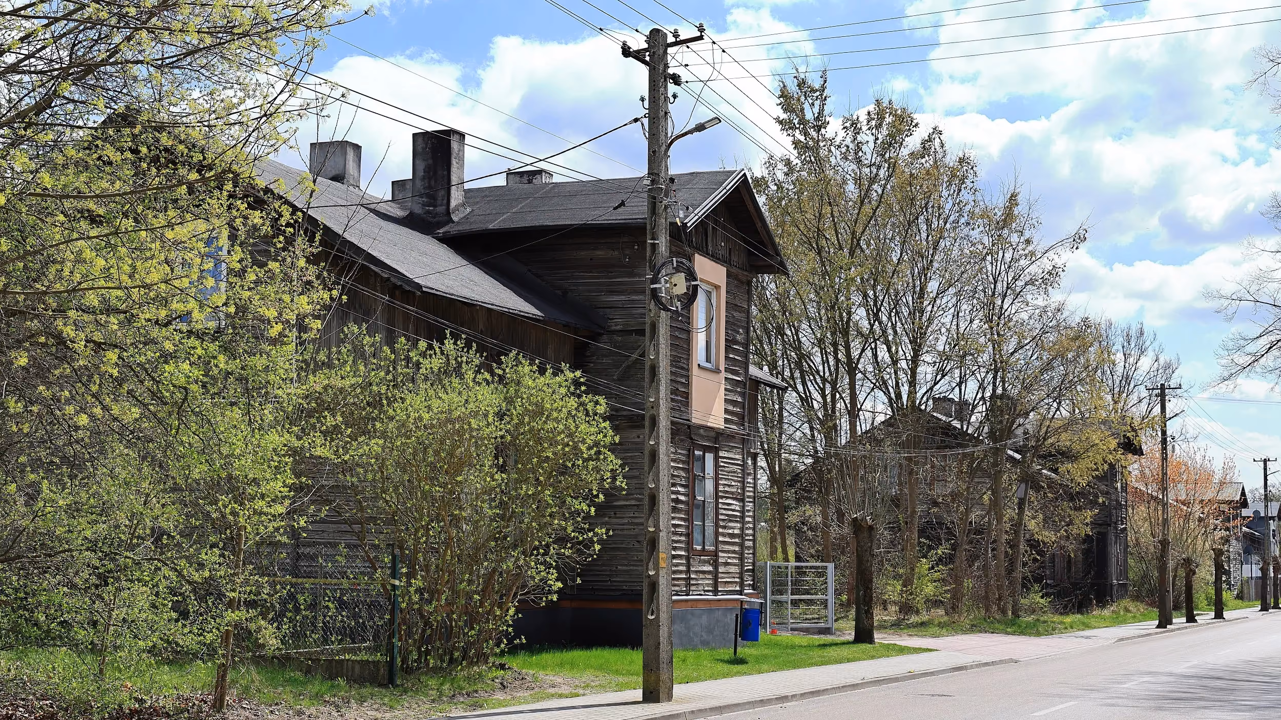 Widok boczny zabytkowego drewnianego budynku przy ulicy. Wokół rosną drzewa i krzewy, a przed obiektem przebiega chodnik oraz linie energetyczne.