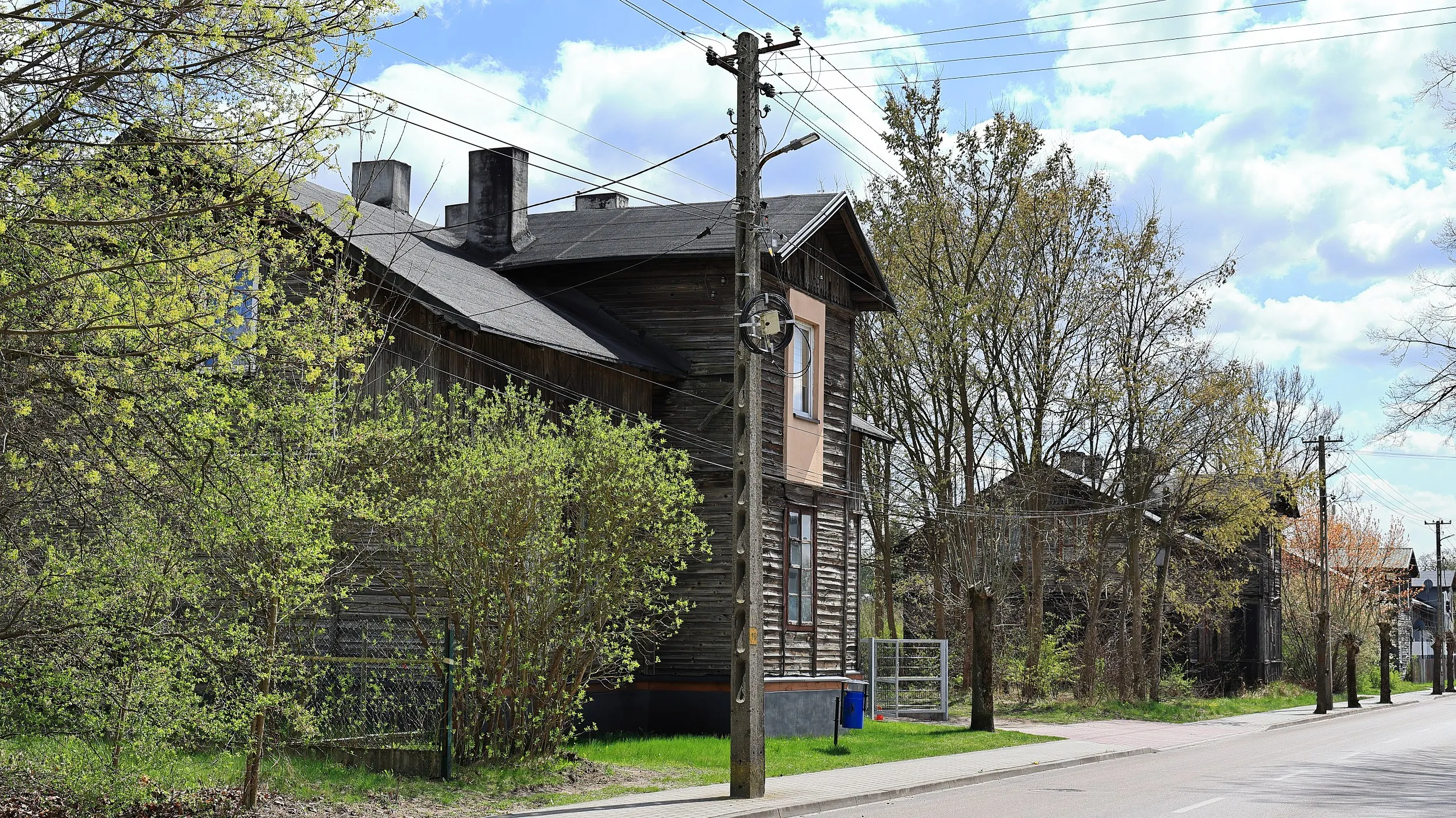 Widok boczny zabytkowego drewnianego budynku przy ulicy. Wokół rosną drzewa i krzewy, a przed obiektem przebiega chodnik oraz linie energetyczne.