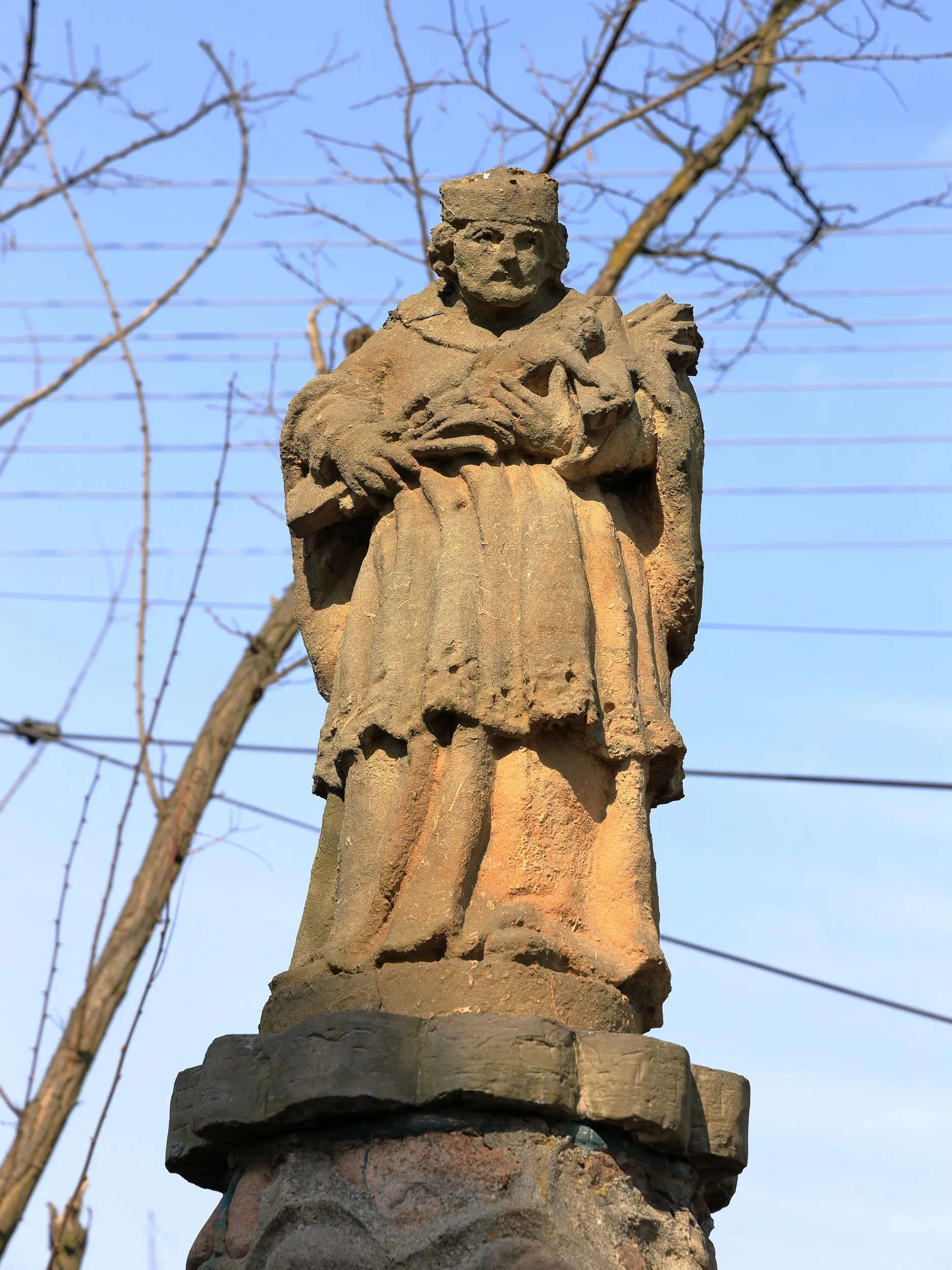 Kamienna figura świętego ustawiona na ozdobnym postumencie. W tle widoczne są gałęzie drzew i niebo.