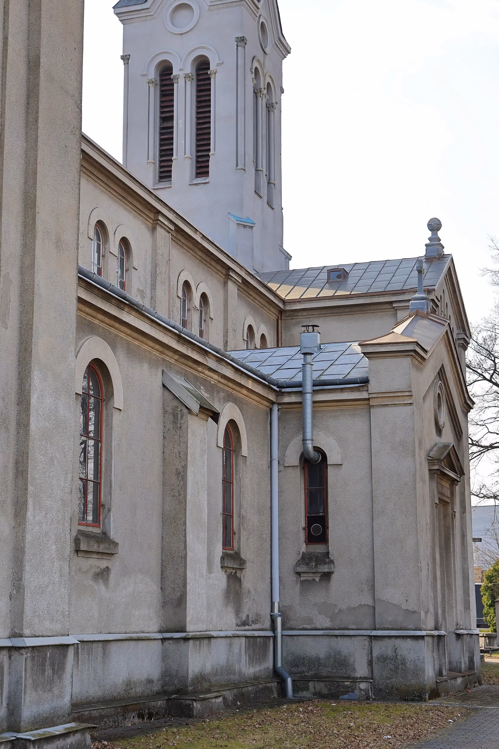 Boczne ujęcie jasnego kościoła z wysoką wieżą zakończoną strzelistym hełmem. Wzdłuż budynku biegnie brukowana alejka.
