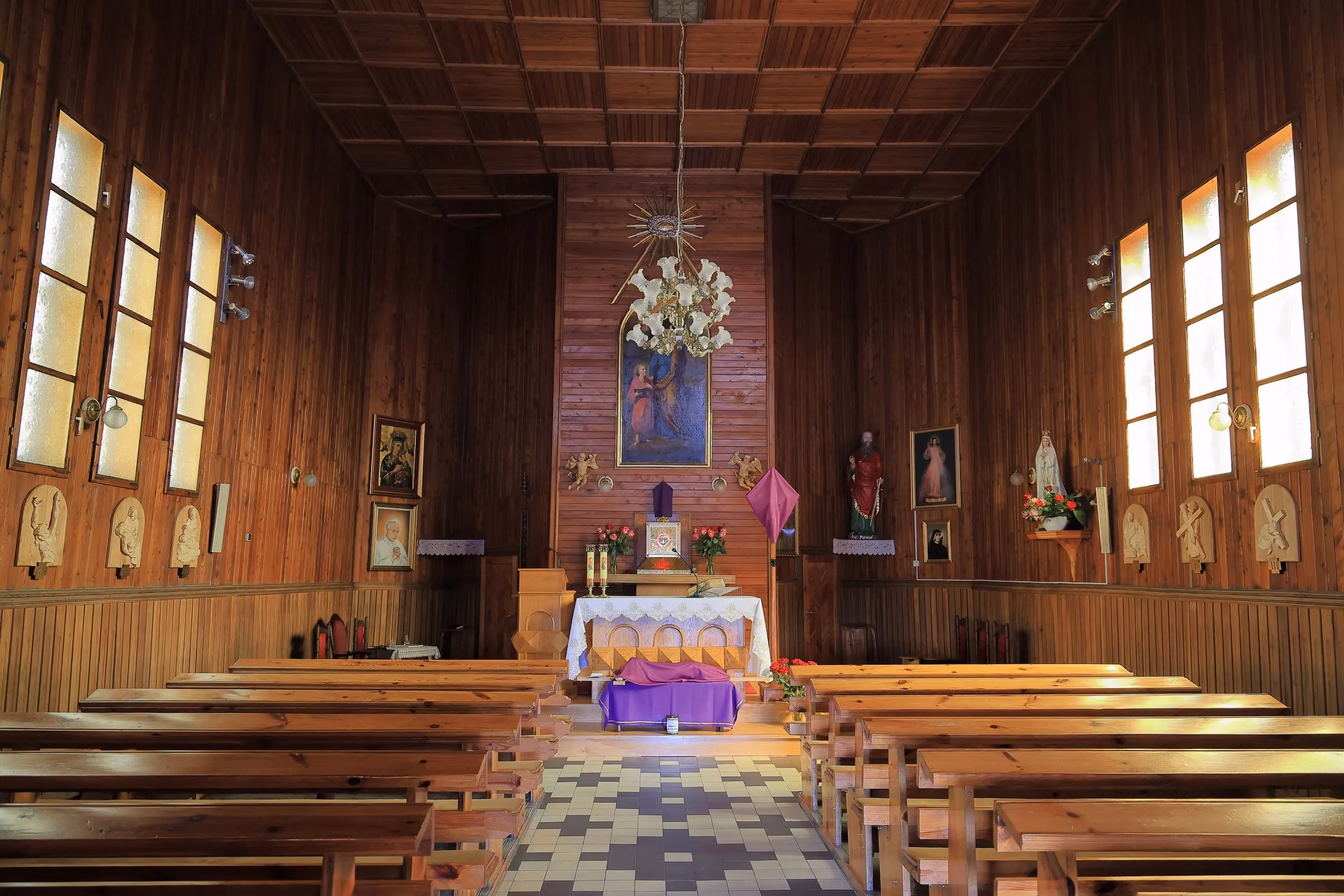 Wnętrze drewnianego kościoła z rzędami ławek prowadzących do ołtarza. Ściany zdobią obrazy religijne, rzeźby oraz drewniana boazeria.