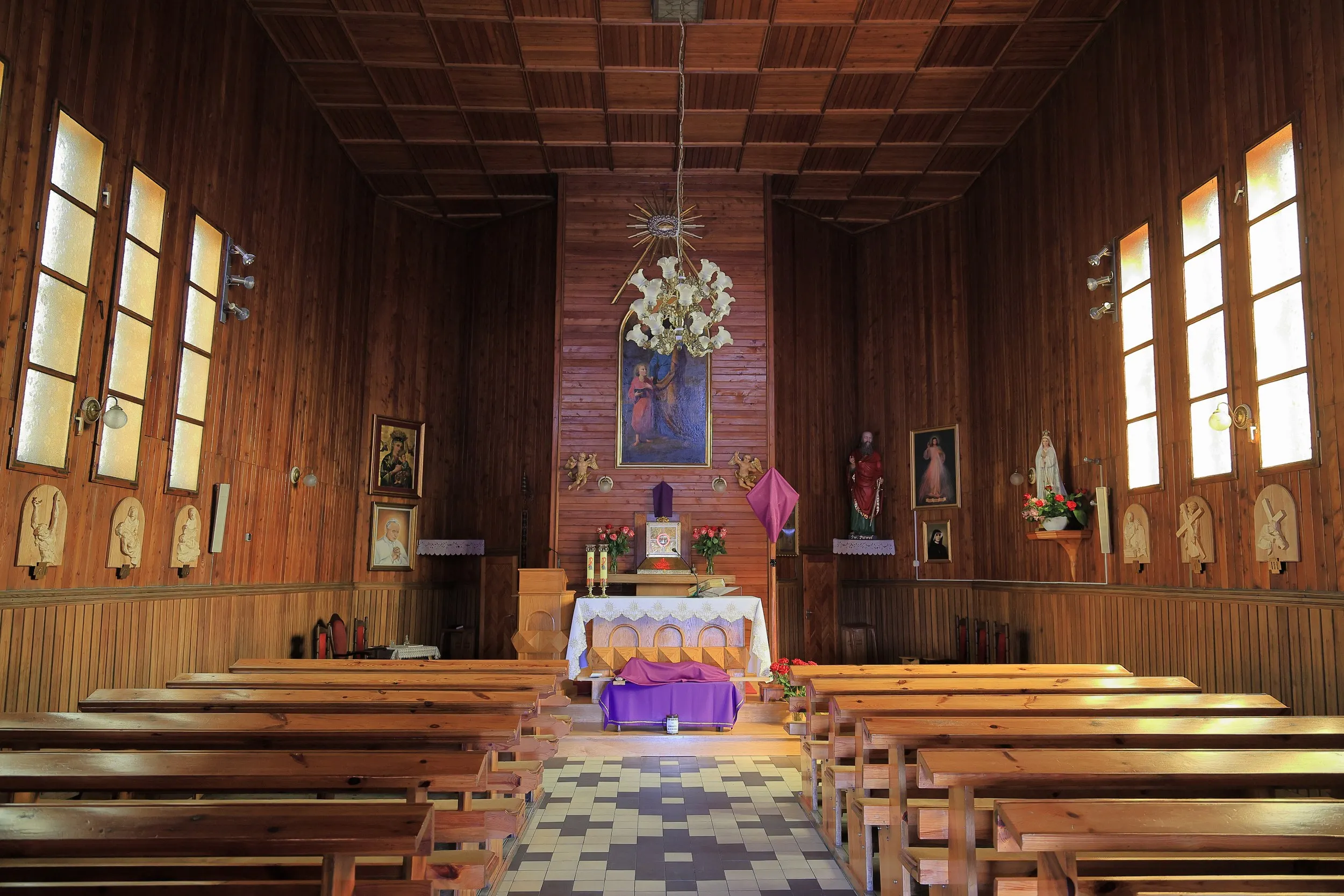 Wnętrze drewnianego kościoła z rzędami ławek prowadzących do ołtarza. Ściany zdobią obrazy religijne, rzeźby oraz drewniana boazeria.