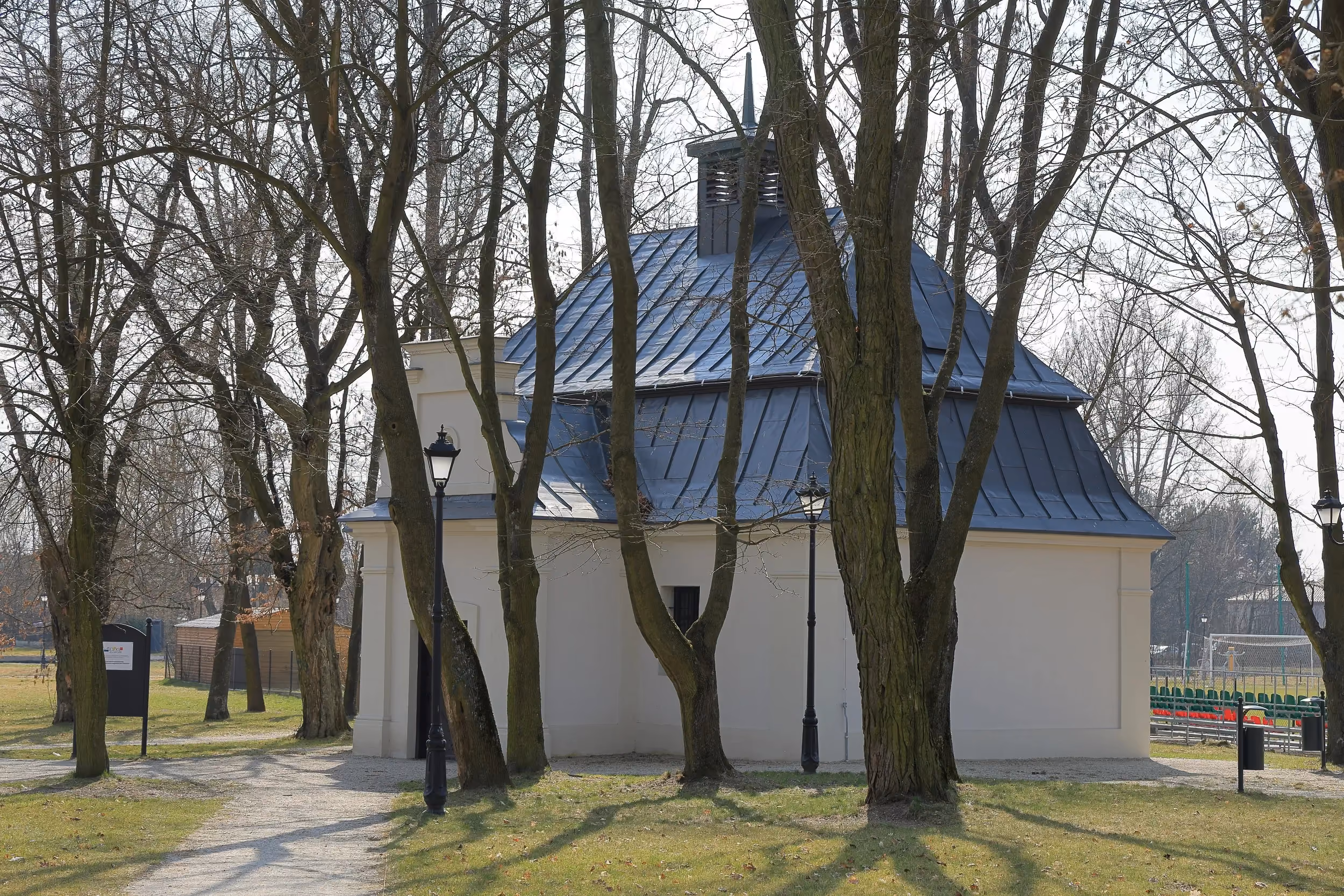 Lamus w Gorzędowie widziany od strony bocznej. Jasny budynek z ciemnym dachem otaczają drzewa i parkowe alejki.