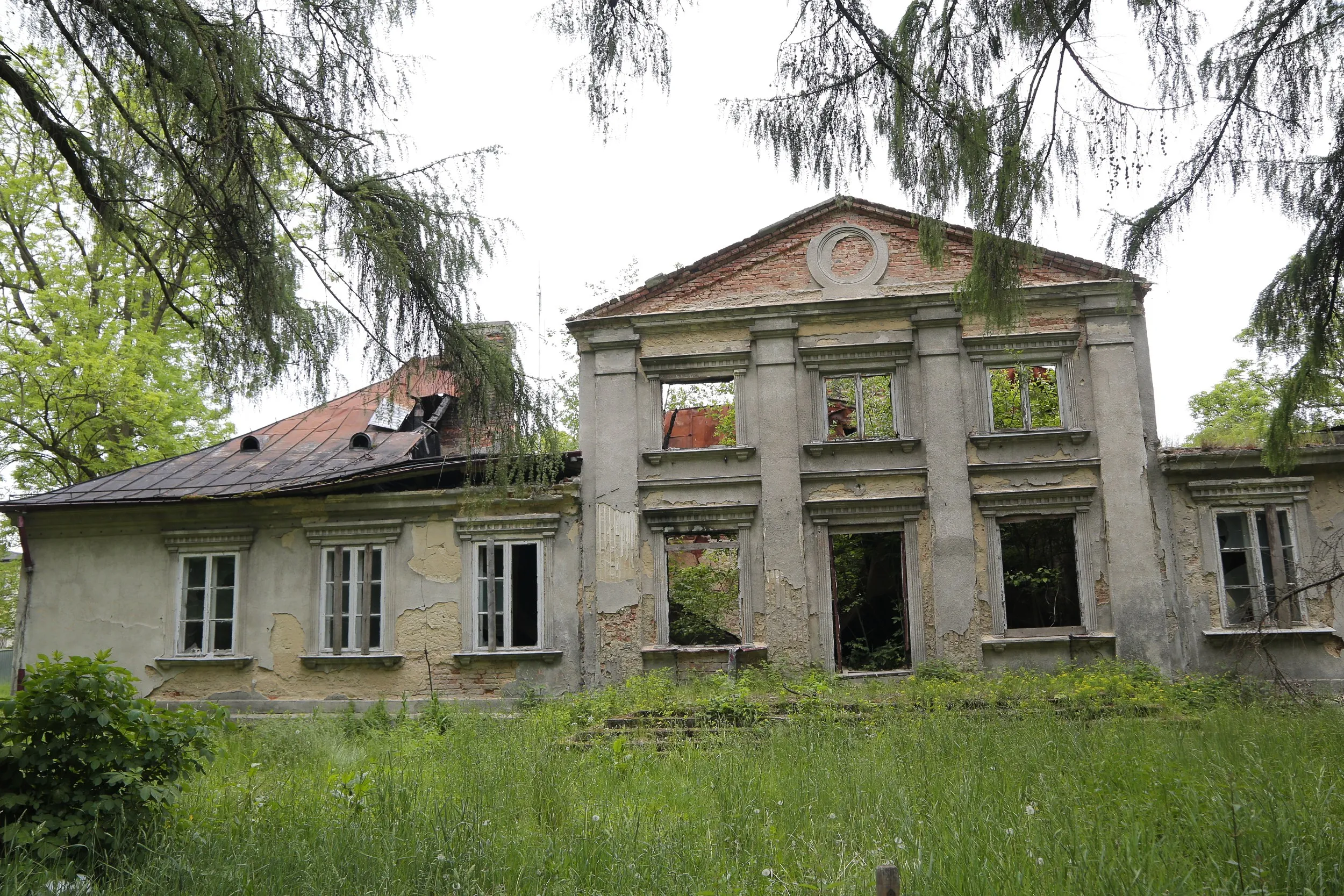 Szeroki widok na ruiny dworu Tymowskich w Kobielach Wielkich. Budynek z uszkodzonym dachem i pustymi oknami otaczają drzewa oraz wysoka trawa.