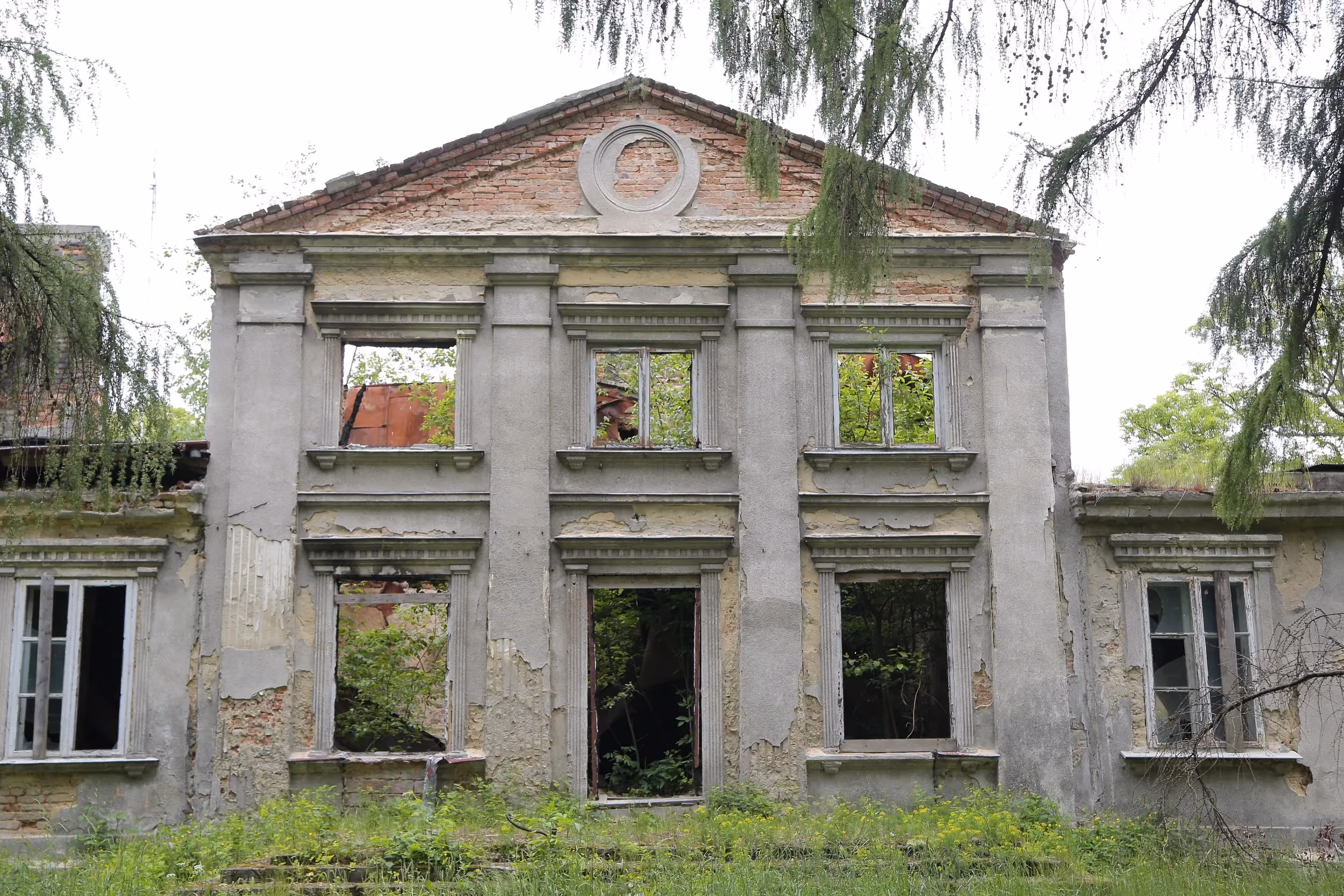 Frontowa elewacja ruin dworu Tymowskich w Kobielach Wielkich. Zniszczony budynek z pustymi oknami otacza wysoka trawa i samosiewna roślinność.