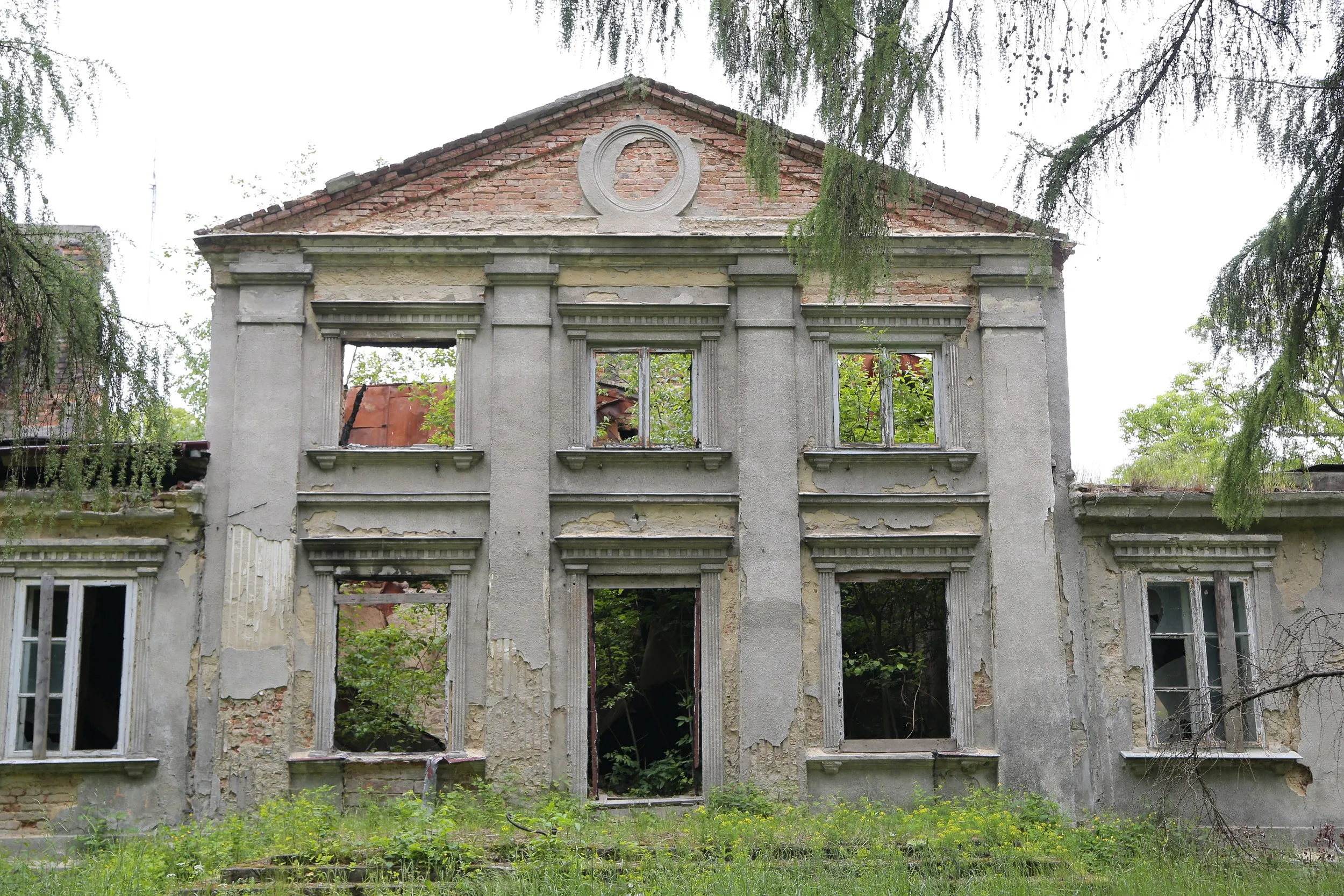 Frontowa elewacja ruin dworu Tymowskich w Kobielach Wielkich. Zniszczony budynek z pustymi oknami otacza wysoka trawa i samosiewna roślinność.