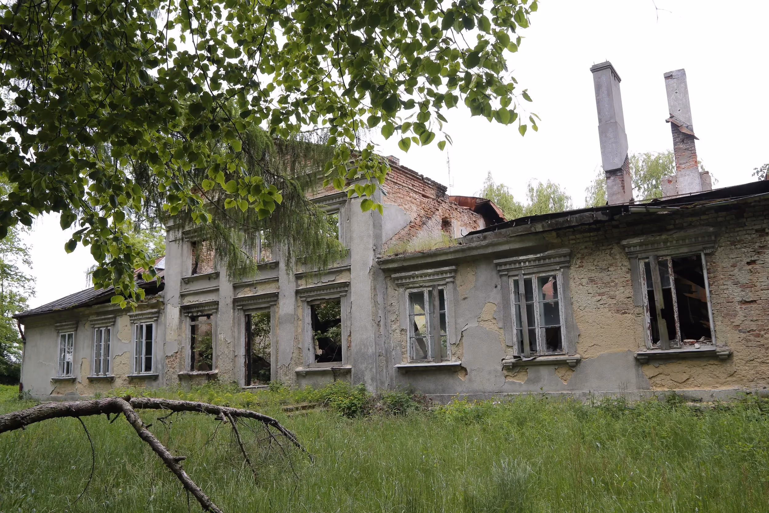 Widok dworu Tymowskich z boku. Opuszczony budynek z uszkodzonym dachem i wybitymi oknami stoi pośród gęstej zieleni.