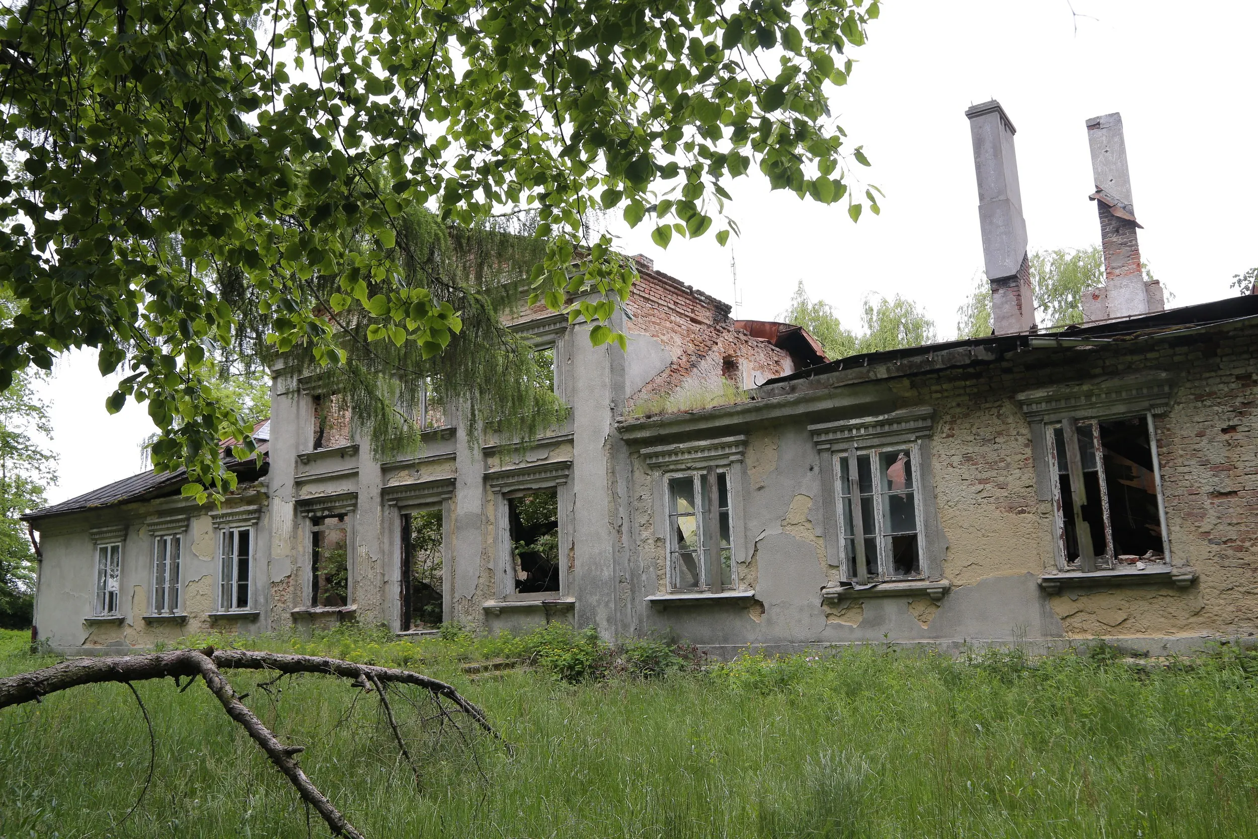 Widok dworu Tymowskich z boku. Opuszczony budynek z uszkodzonym dachem i wybitymi oknami stoi pośród gęstej zieleni.