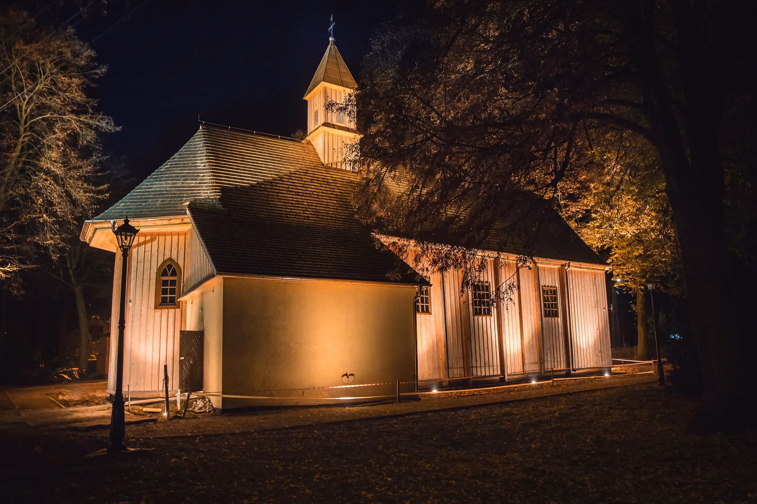 Kościół w Rzejowicach nocą, oświetlona drewniana świątynia otoczona drzewami i parkowym terenem.