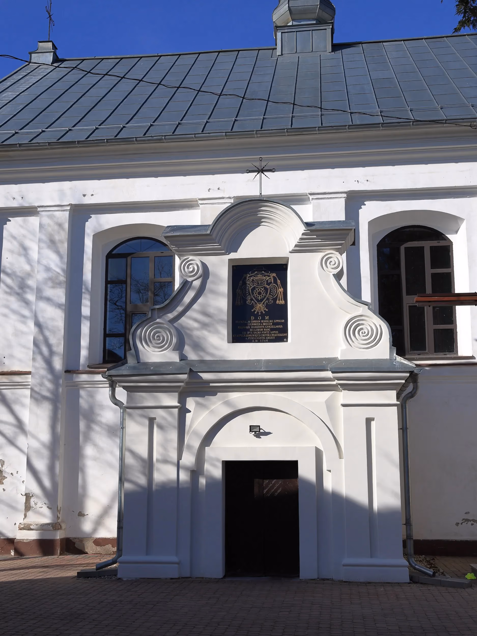 Wejście na teren kościoła w Dmeninie z ozdobną bramą oraz widoczną fasadą świątyni w tle.