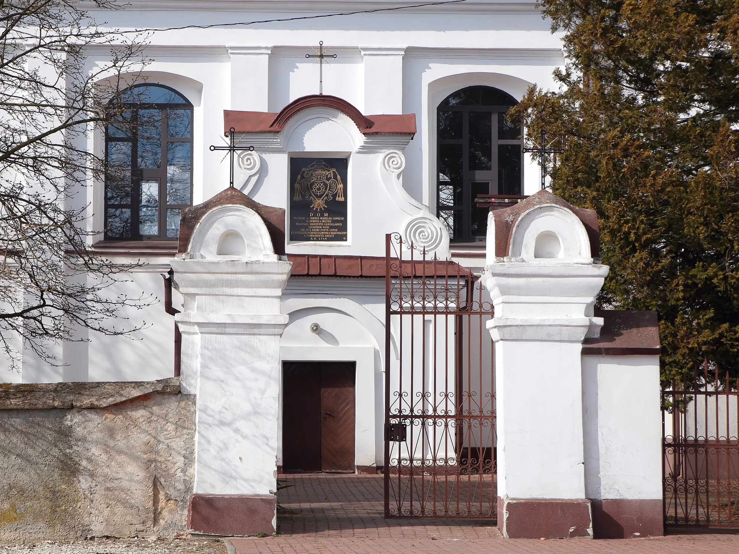 Barokowy portal wejściowy kościoła w Dmeninie z tablicą fundacyjną umieszczoną nad głównym wejściem.