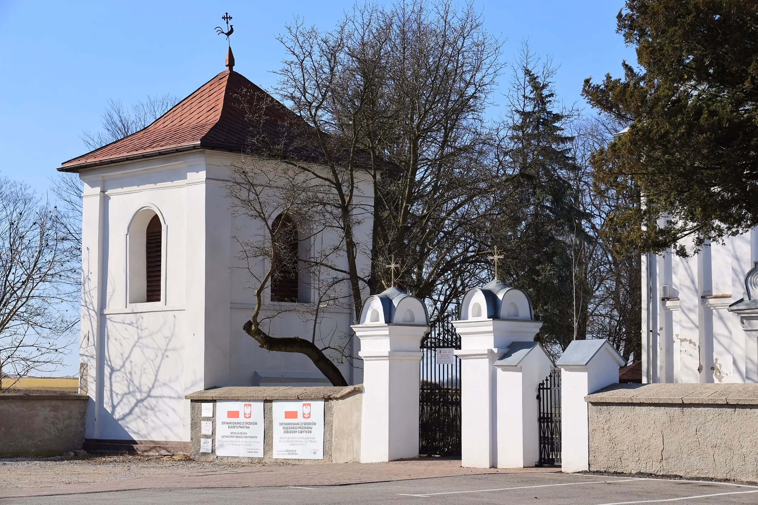 Dzwonnica i brama wejściowa na teren kościoła w Dmeninie, widoczne na tle drzew i zabudowań parafialnych.
