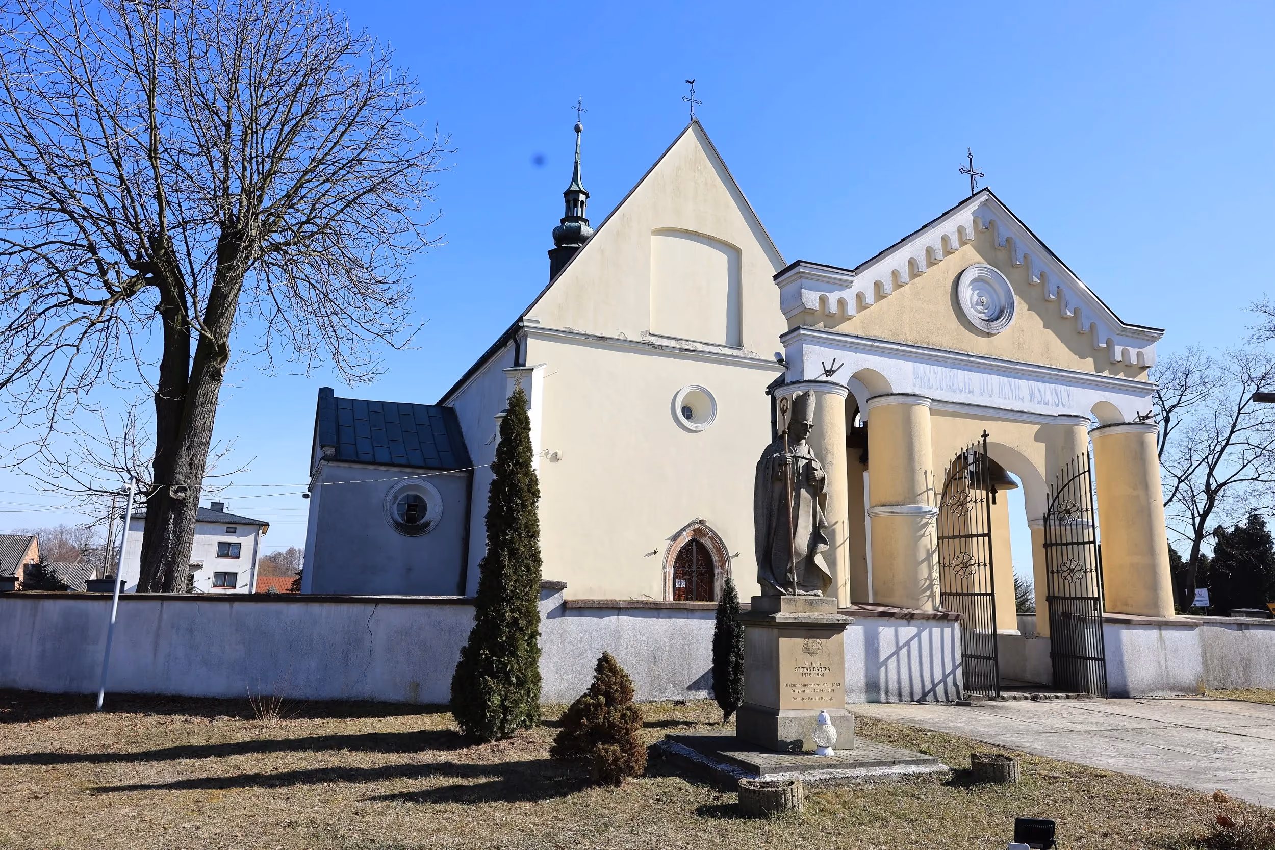 Boczny widok kościoła pw. św. Marcina w Kodrębie z bramą wejściową i figurą świętego na postumencie. Budowlę otacza mur i zieleń.