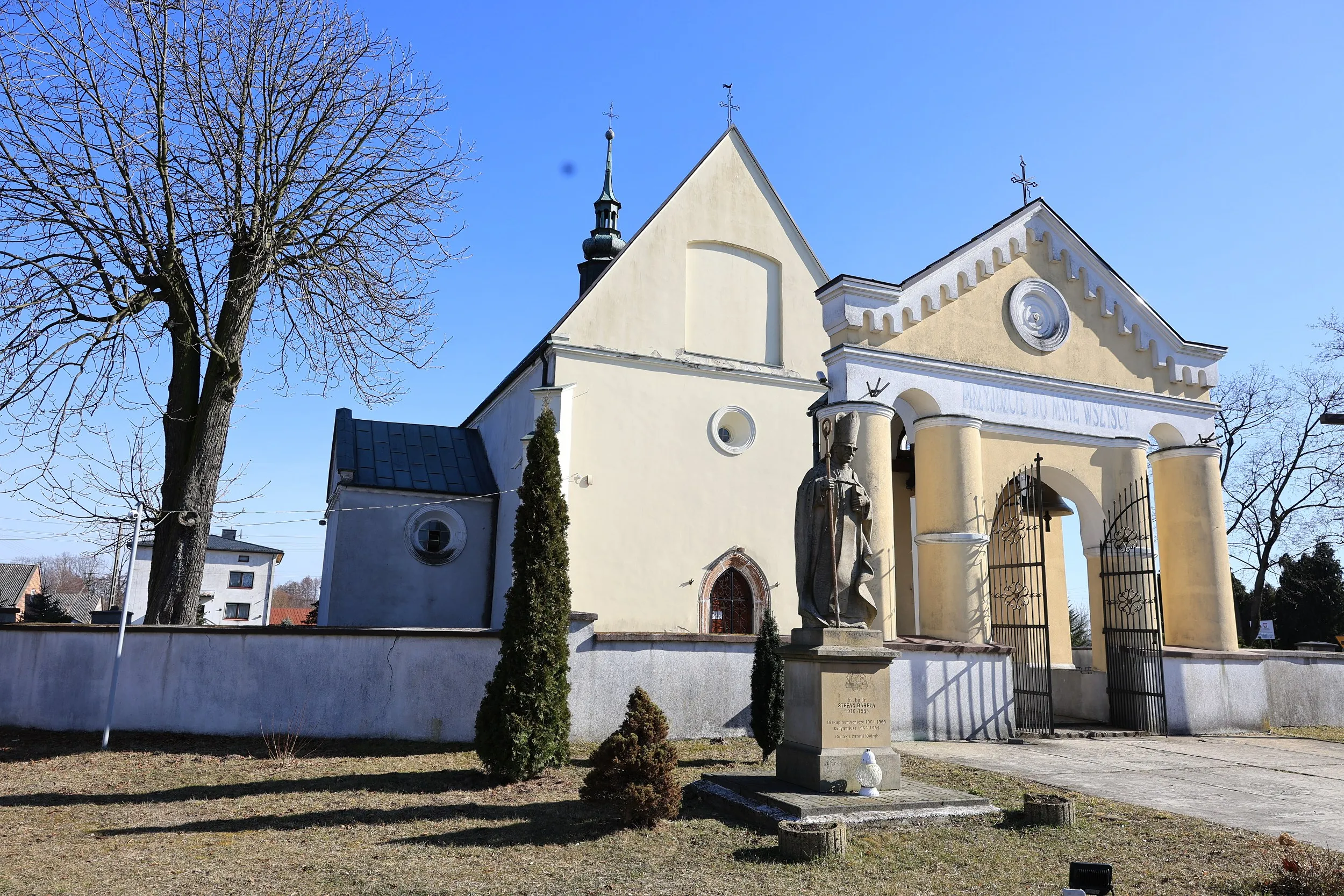 Boczny widok kościoła pw. św. Marcina w Kodrębie z bramą wejściową i figurą świętego na postumencie. Budowlę otacza mur i zieleń.