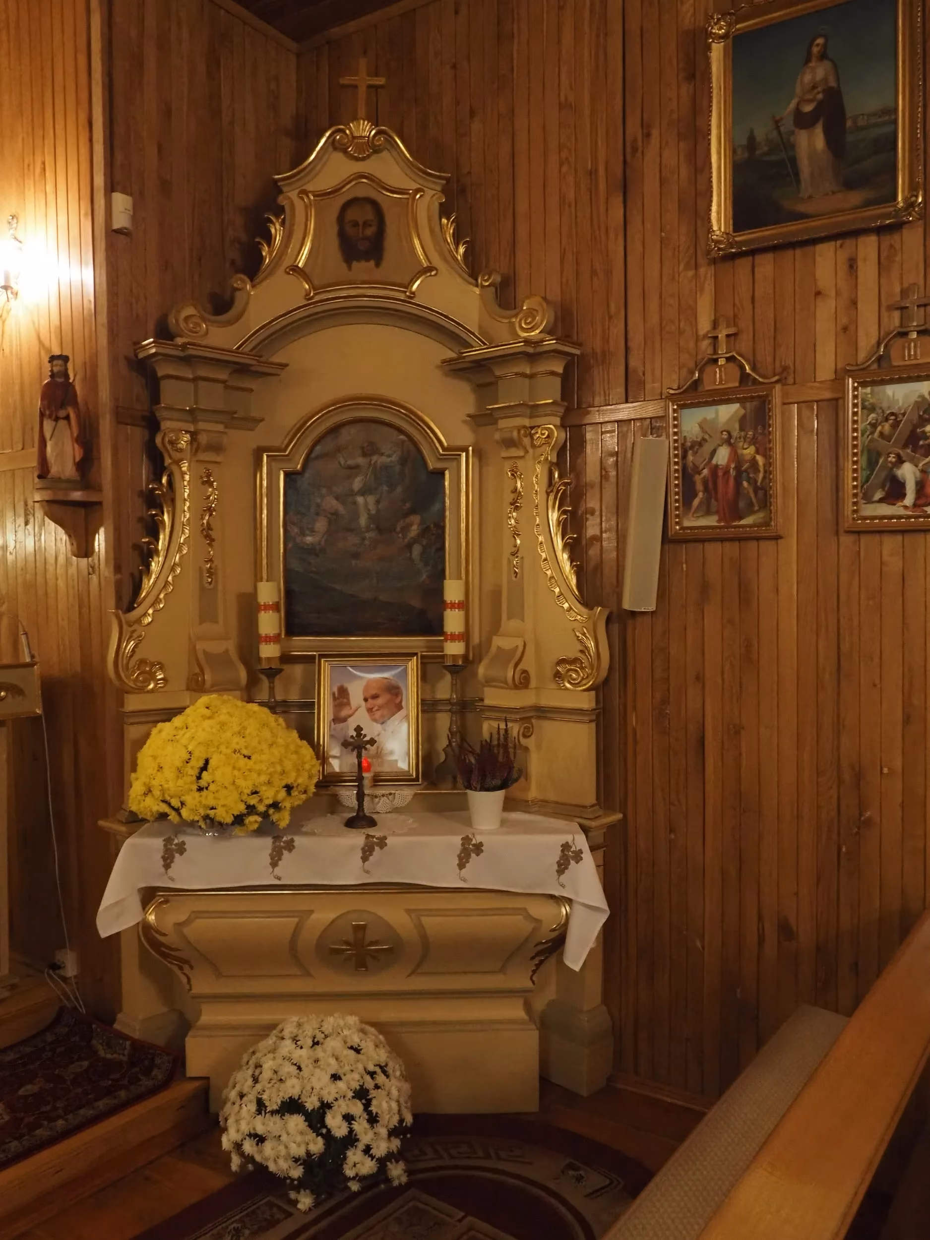 Boczny ołtarz we wnętrzu kościoła w Bugaju Zakrzewskim, ozdobiony obrazami, kwiatami i fotografią św. Jana Pawła II.
