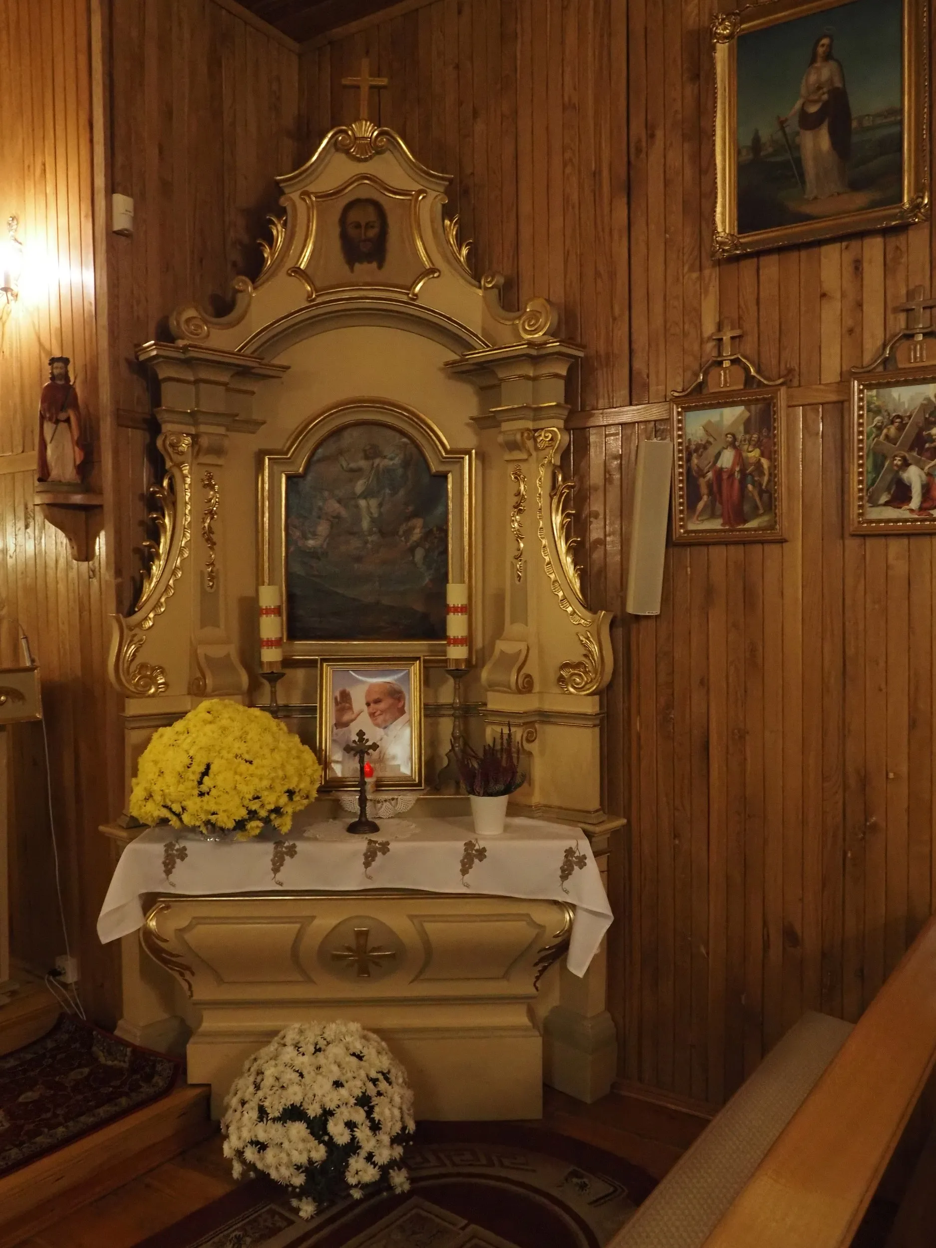 Boczny ołtarz we wnętrzu kościoła w Bugaju Zakrzewskim, ozdobiony obrazami, kwiatami i fotografią św. Jana Pawła II.