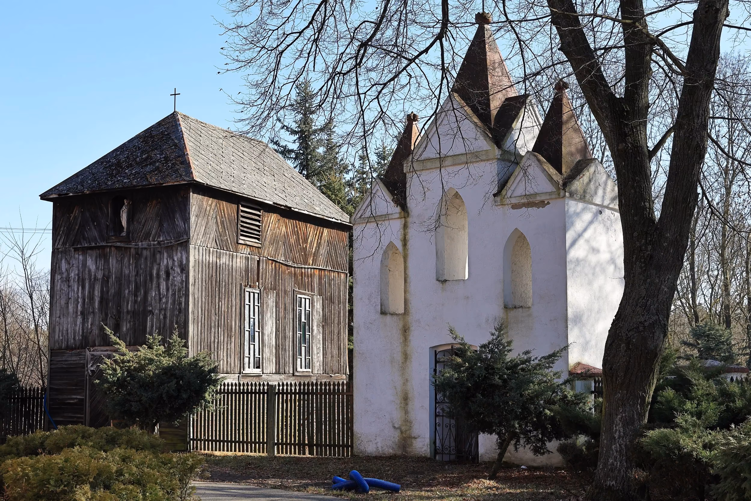 Drewniana dzwonnica i zabytkowa murowana dzwonnica przy kościele w Krępie. Obiekty otoczone są drzewami i ogrodzeniem na terenie przykościelnym.