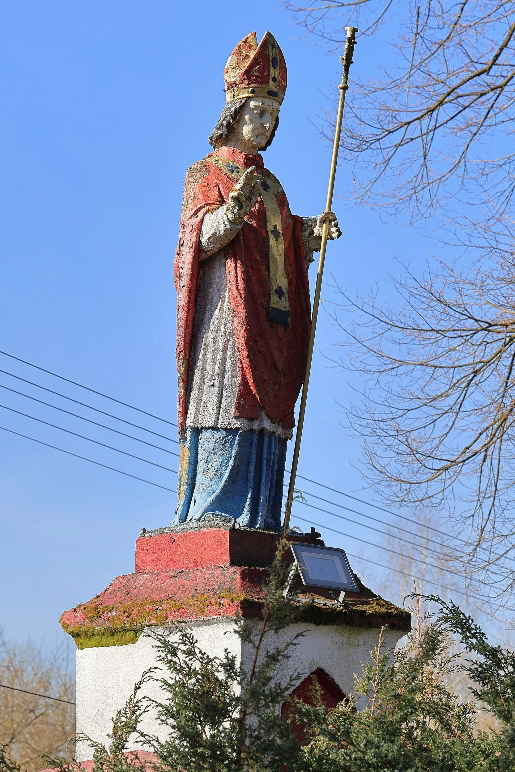 Figura świętego biskupa przy kościele w Krępie. Rzeźba ustawiona na postumencie przedstawia postać w szatach liturgicznych z pastorałem.