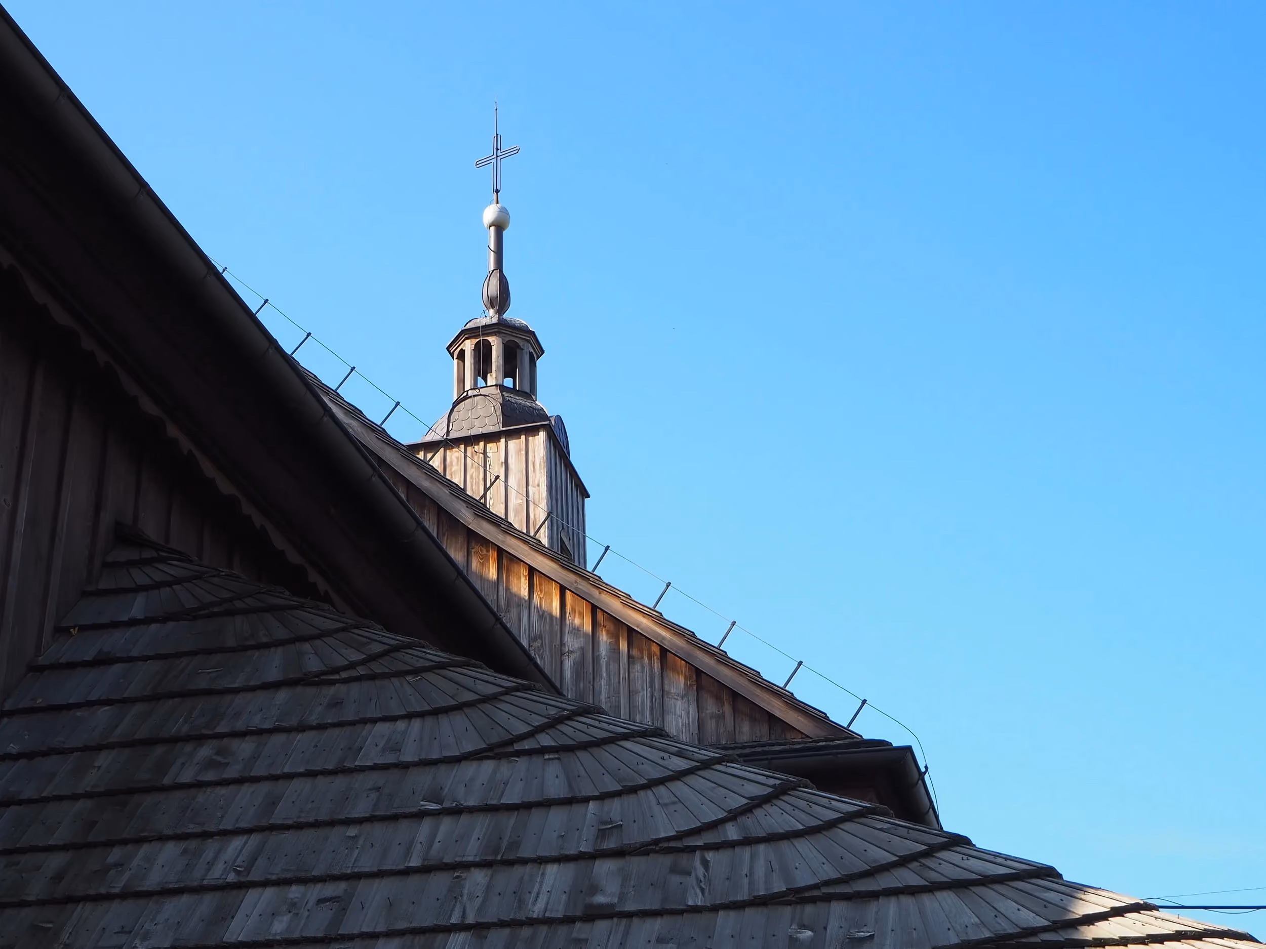 Fragment dachu i wieży drewnianego kościoła w Lgocie Wielkiej. Nad budynkiem góruje krzyż na tle bezchmurnego nieba.