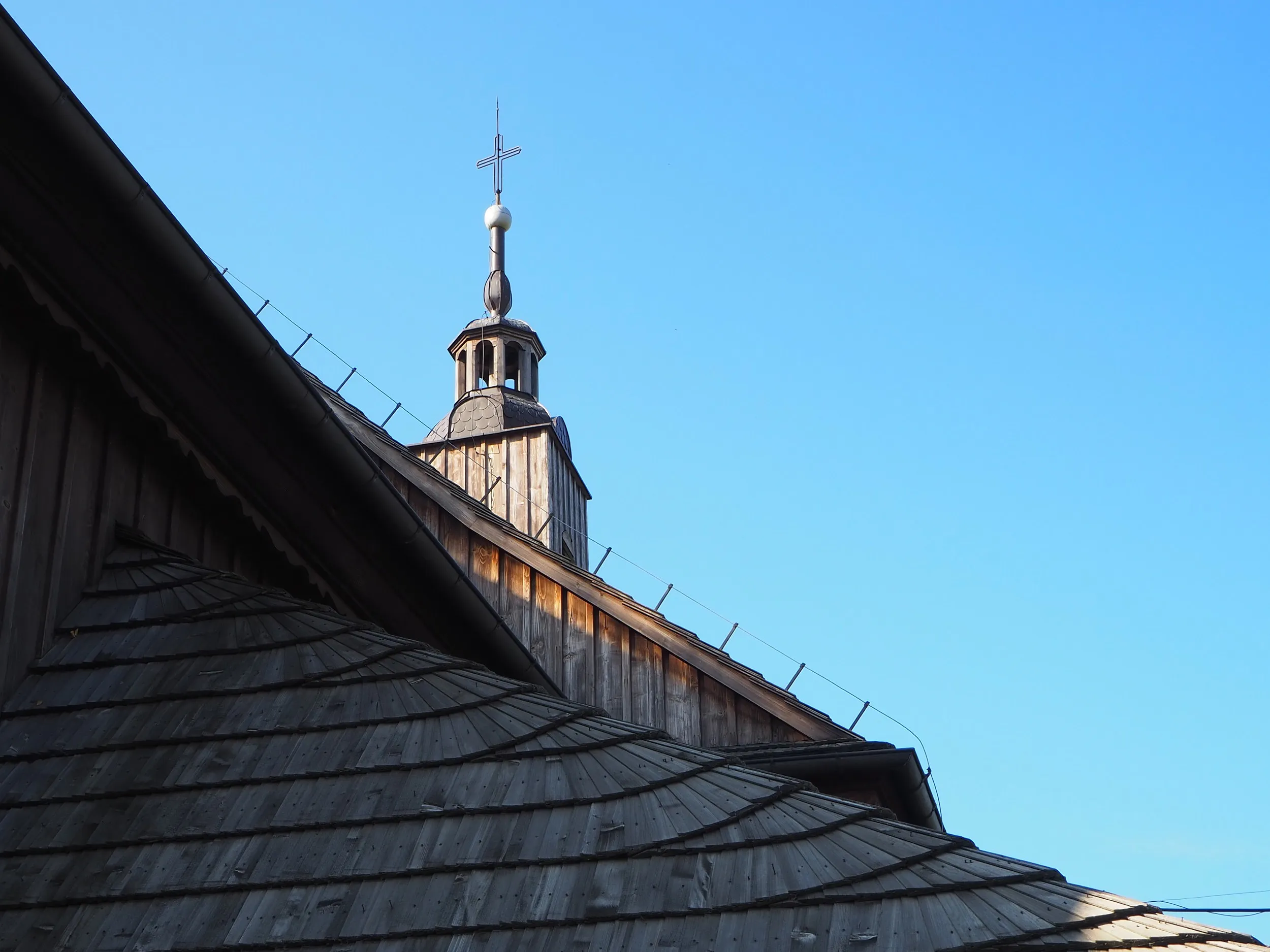 Fragment dachu i wieży drewnianego kościoła w Lgocie Wielkiej. Nad budynkiem góruje krzyż na tle bezchmurnego nieba.