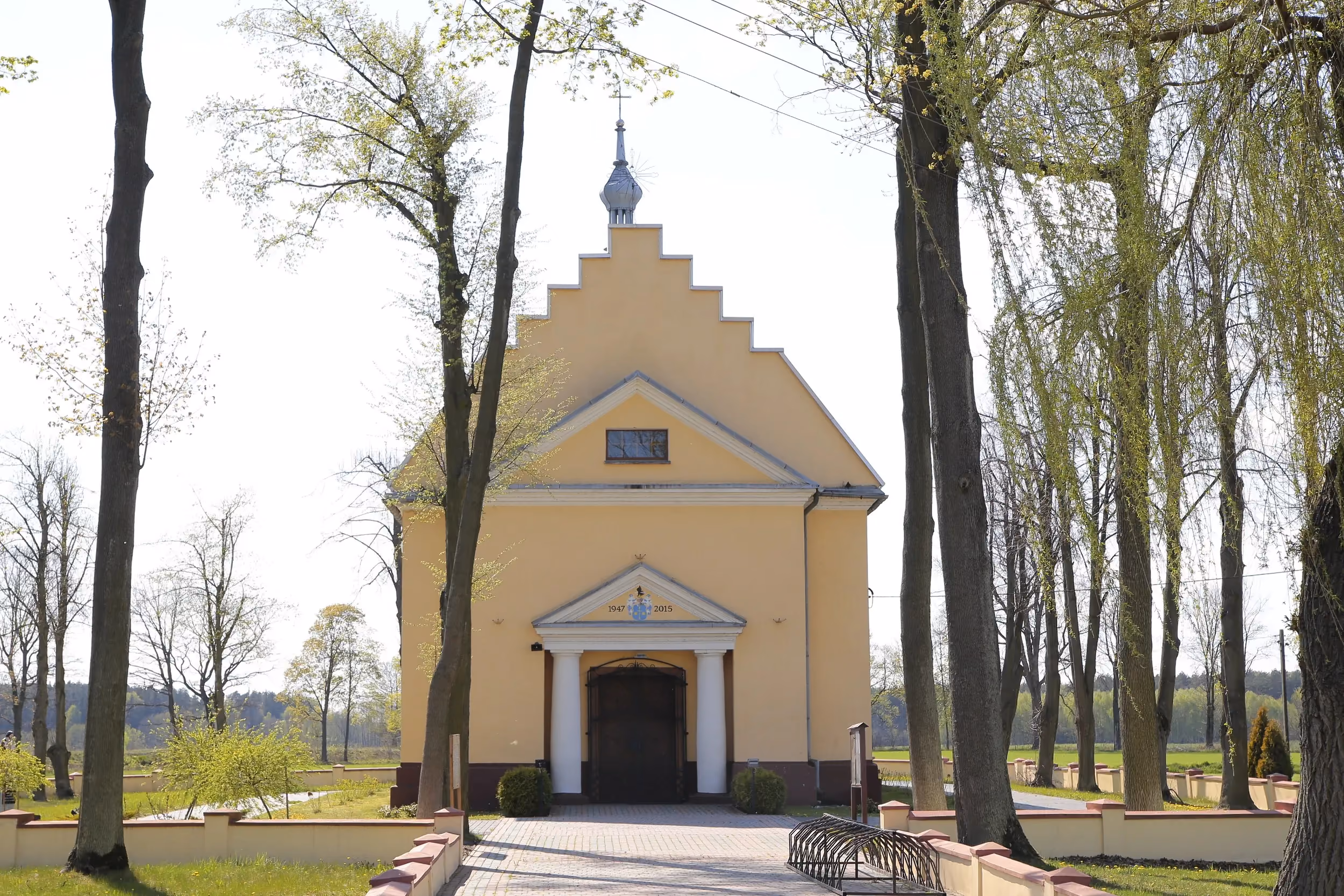 Frontowa elewacja kościoła w Przerębie. Wejście z kolumnowym portykiem prowadzi do świątyni otoczonej wysokimi drzewami i zadbanym terenem przykościelnym.