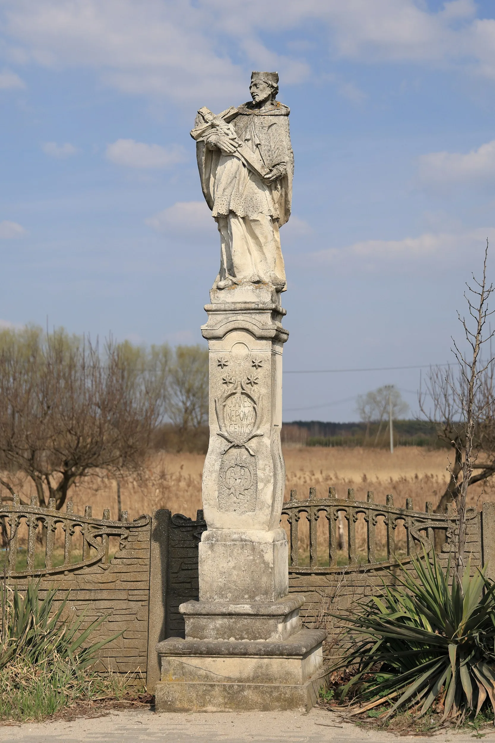 Pełny widok figury św. Jana Nepomucena w Przerębie. Kamienny pomnik stoi na ozdobnym postumencie pośród wiejskiego krajobrazu.