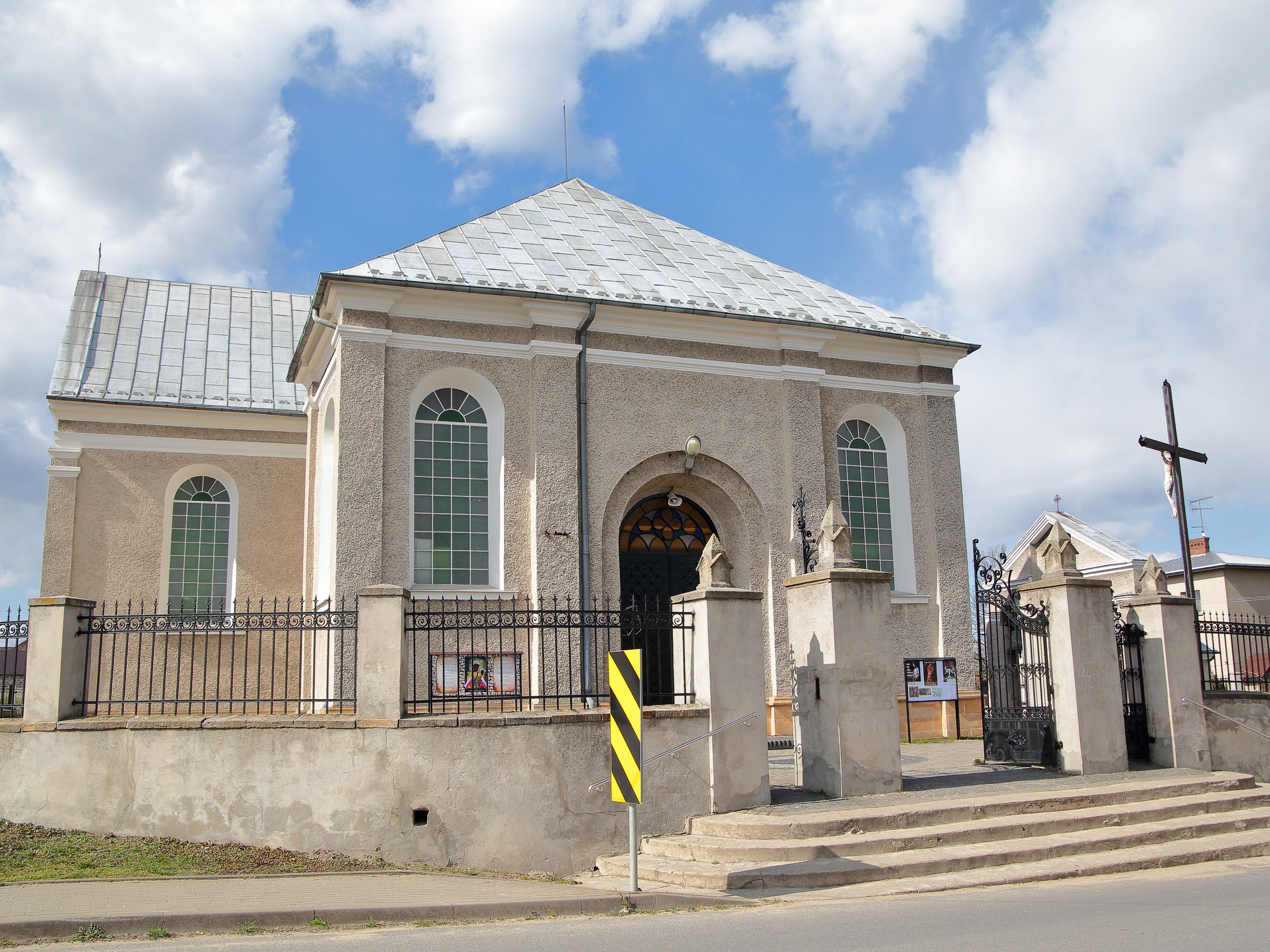 Kościół w Chełmie widziany od strony ulicy. Jasna świątynia z wysokimi oknami, metalowym ogrodzeniem oraz wejściem prowadzącym przez ozdobną bramę.