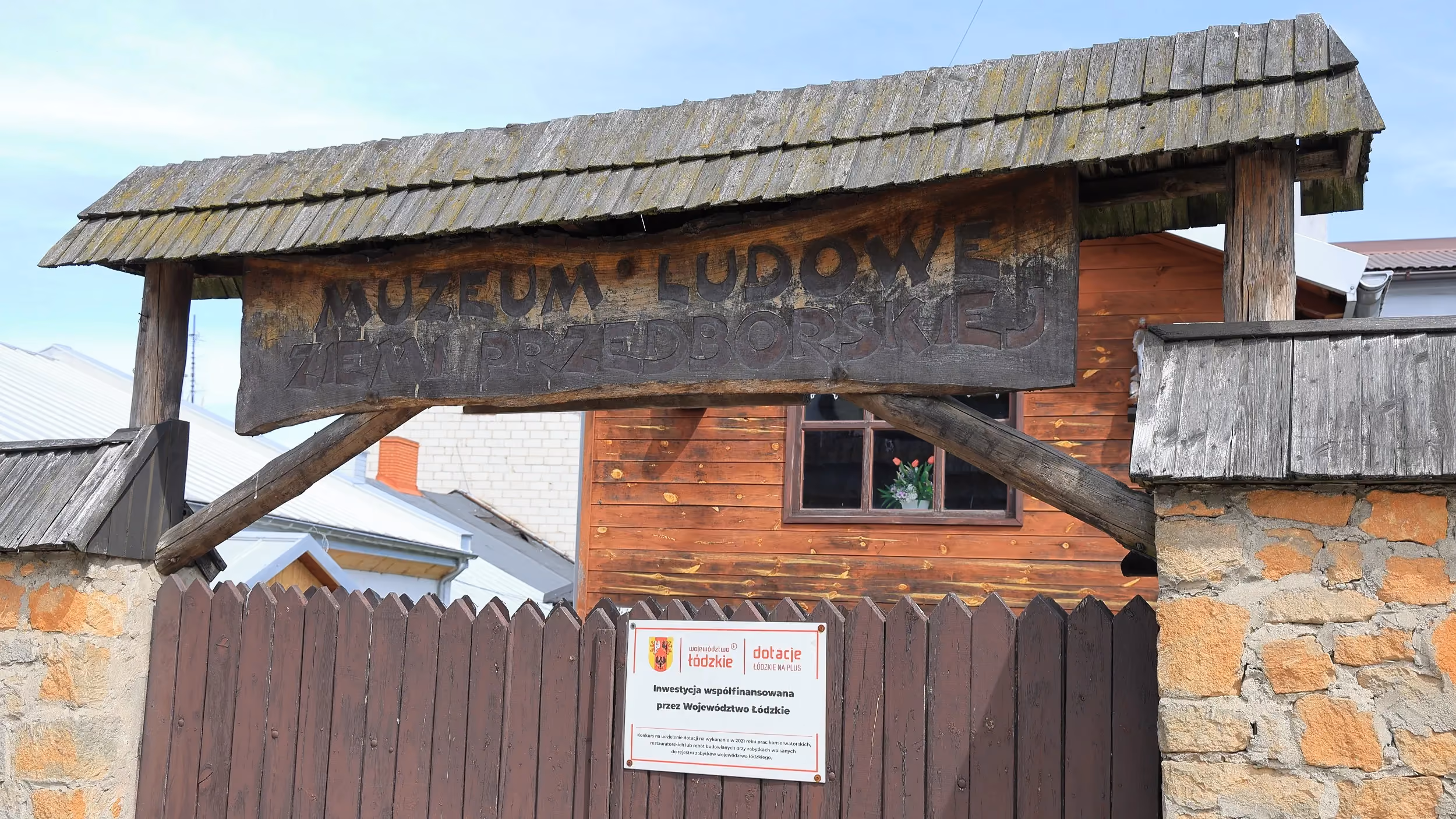 Drewniana brama z szyldem Muzeum Ludowego Ziemi Przedborskiej. Wejście prowadzi na teren muzeum mieszczącego się w zabytkowej dawnej karczmie.