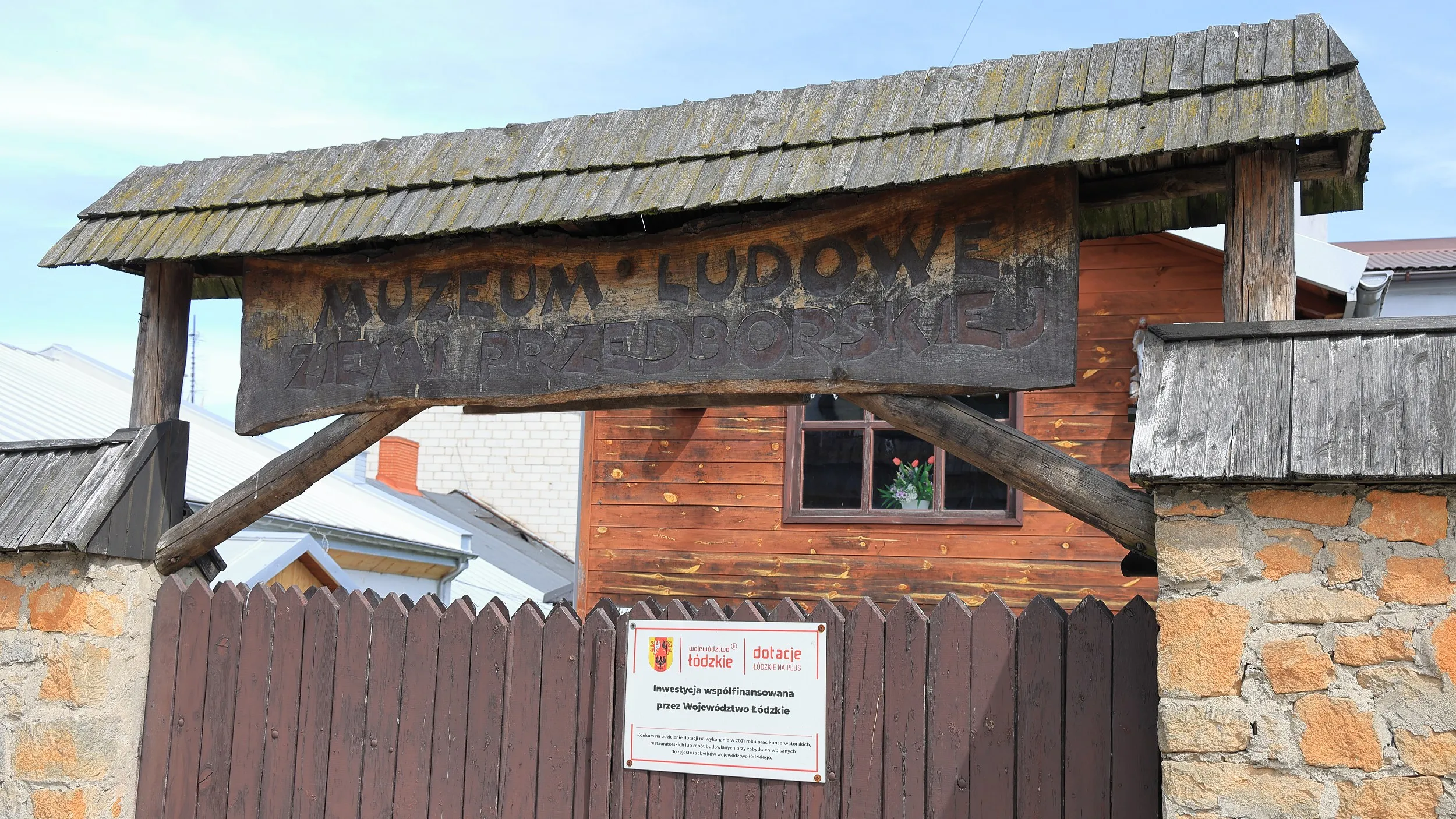 Drewniana brama z szyldem Muzeum Ludowego Ziemi Przedborskiej. Wejście prowadzi na teren muzeum mieszczącego się w zabytkowej dawnej karczmie.