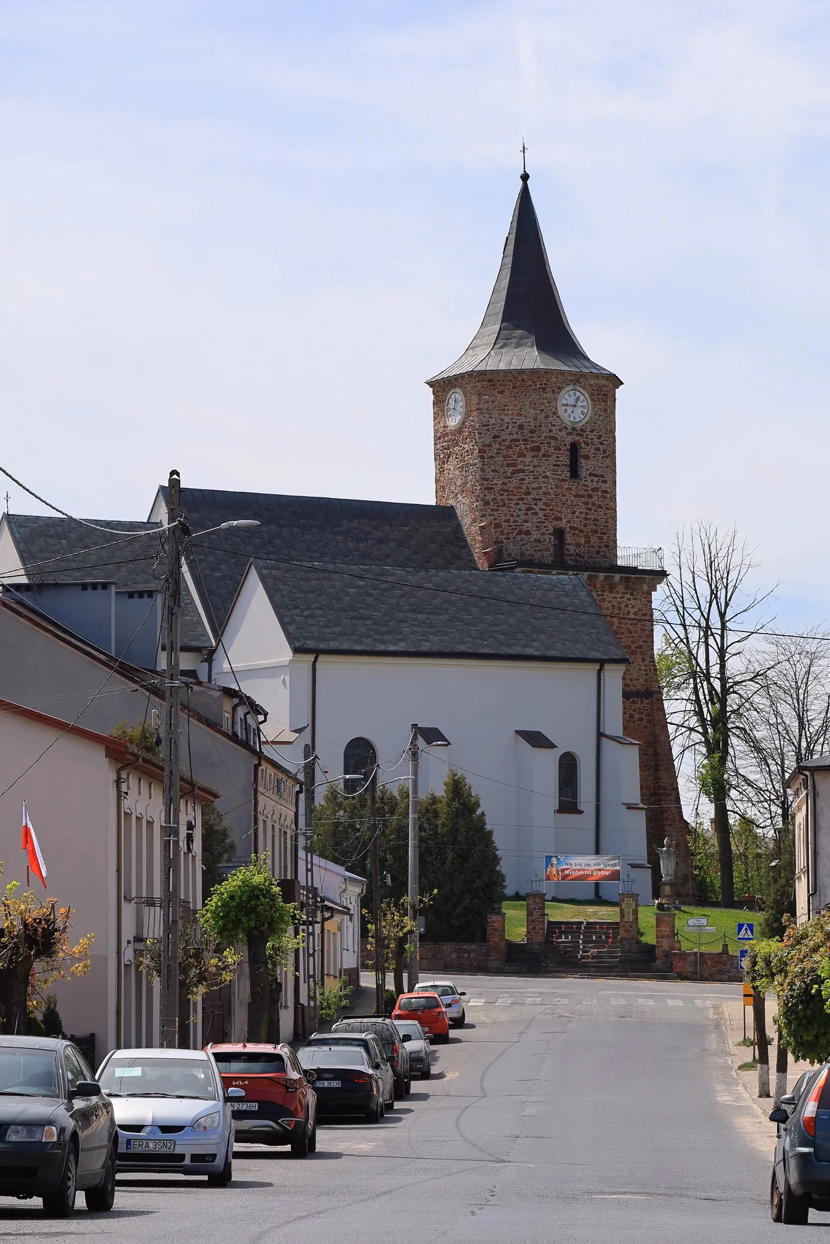 Kościół św. Aleksego górujący nad ulicą w centrum Przedborza. Widoczna ceglana wieża zegarowa i biała bryła świątyni.