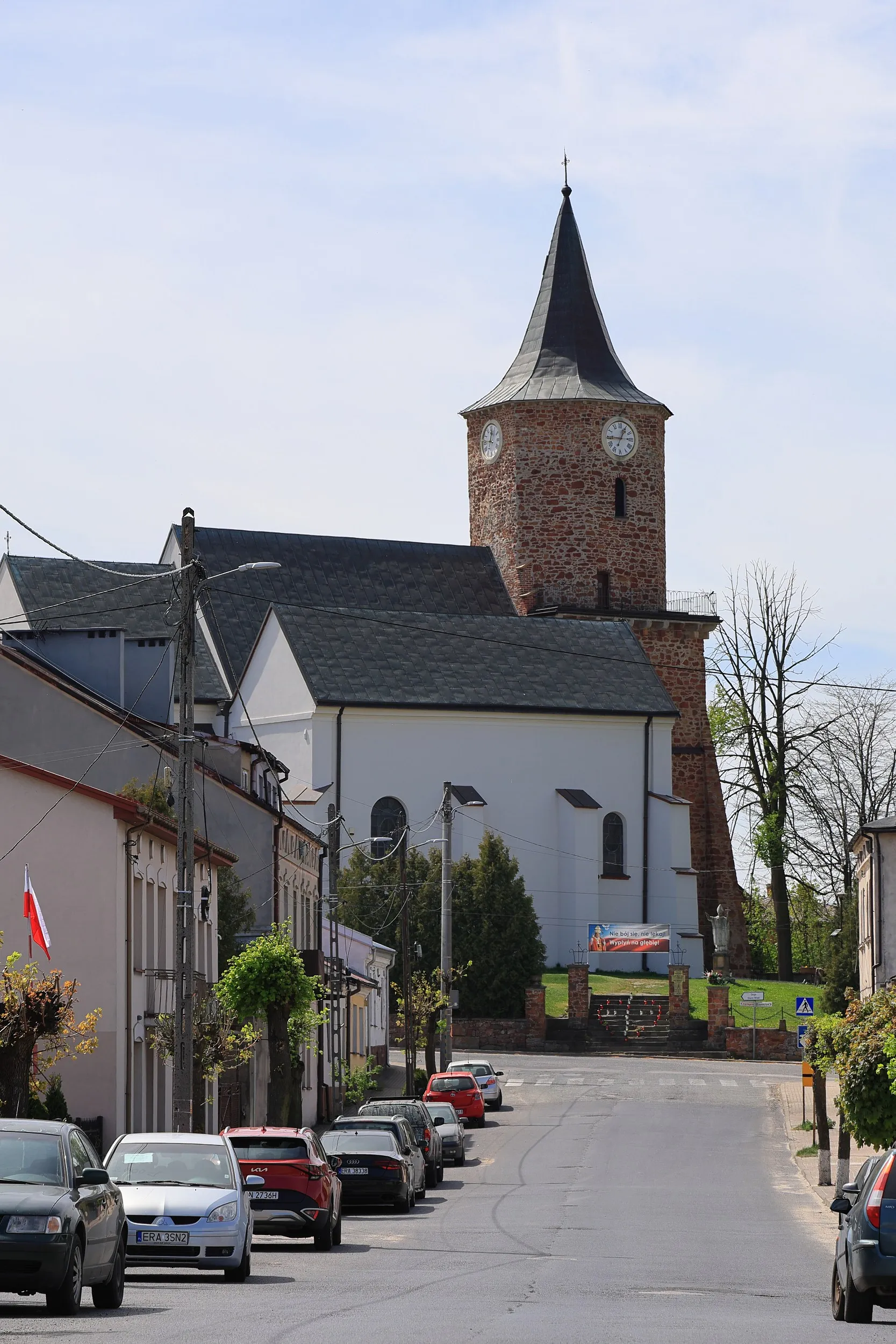 Kościół św. Aleksego górujący nad ulicą w centrum Przedborza. Widoczna ceglana wieża zegarowa i biała bryła świątyni.